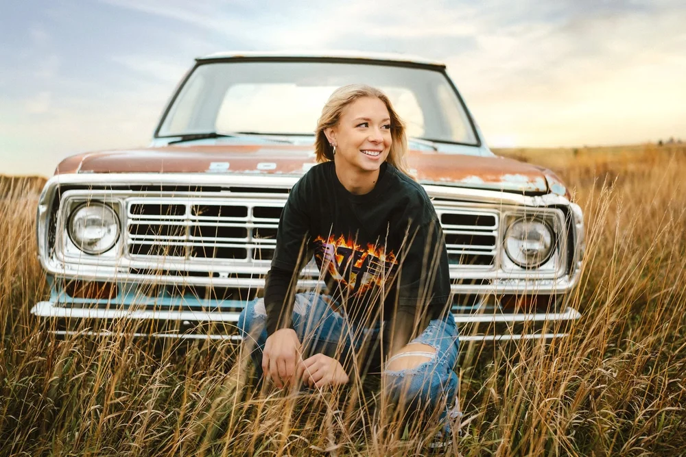 Senior girl kneeling in a field at sunset beside a vintage car