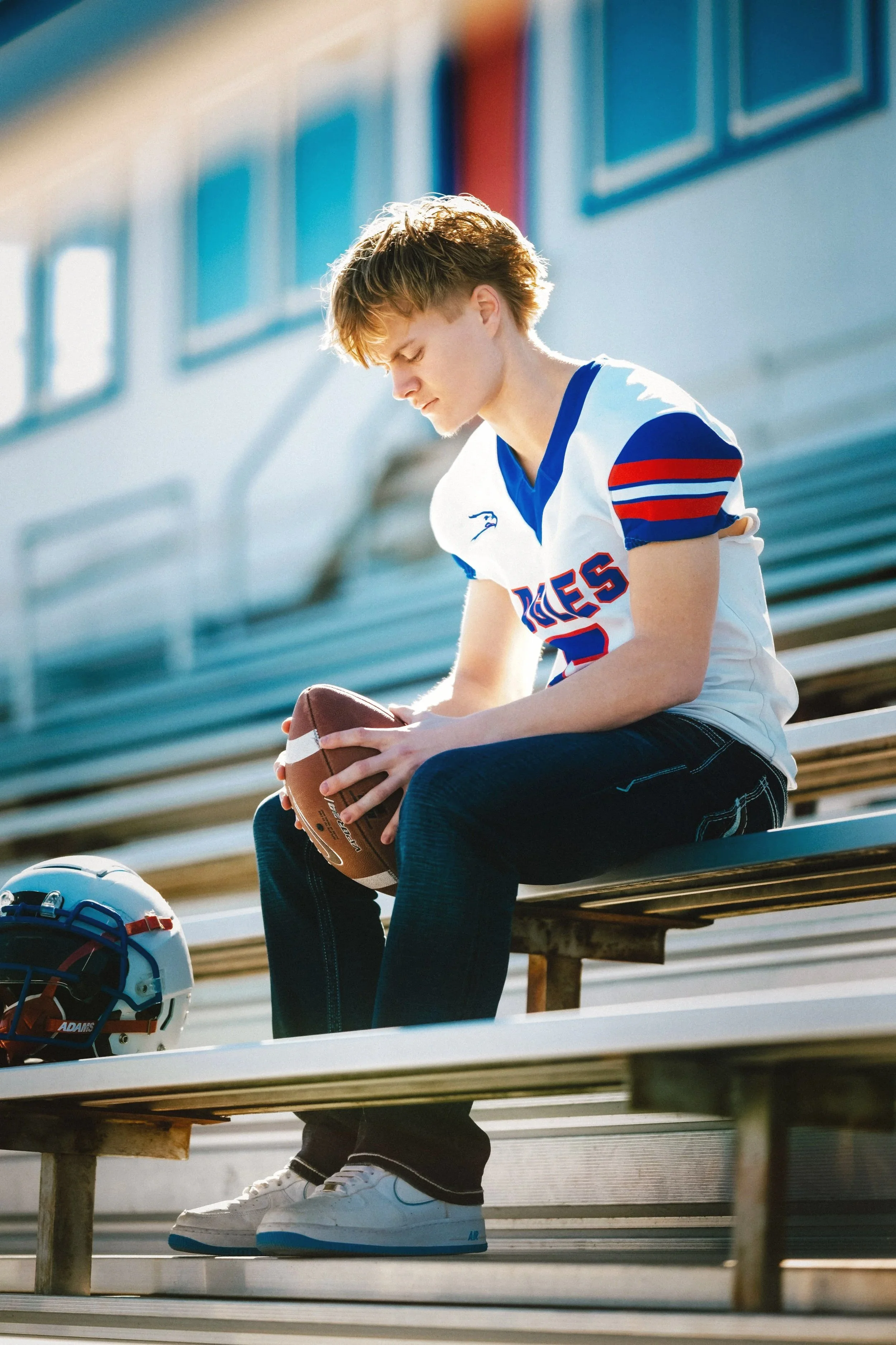 A teenage boy in a football jersey sitting on stadium bleachers, holding a football, with a football helmet on the seat next to him.