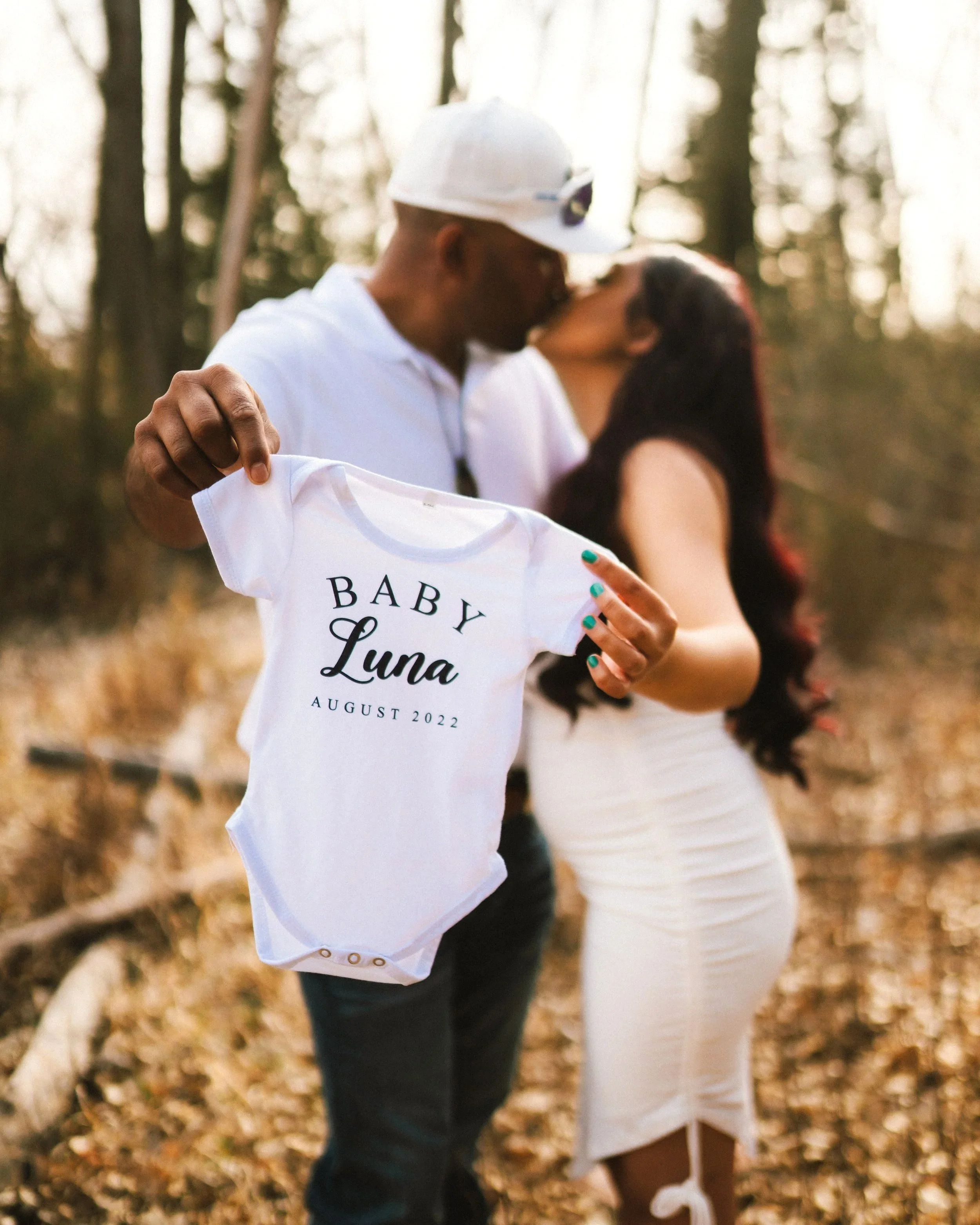 Couple kissing in a forest holding a white baby onesie that reads 'Baby Luna August 2022'.