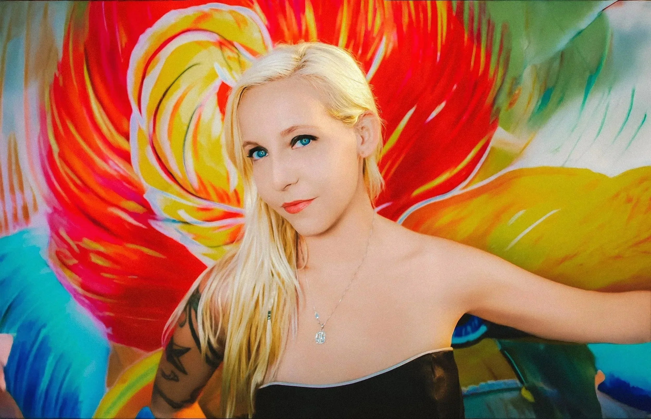 A woman with long blonde hair and blue eyes, wearing a black strapless top and a necklace, taking a selfie in front of a colorful abstract mural with red, yellow, green, and blue swirling patterns.