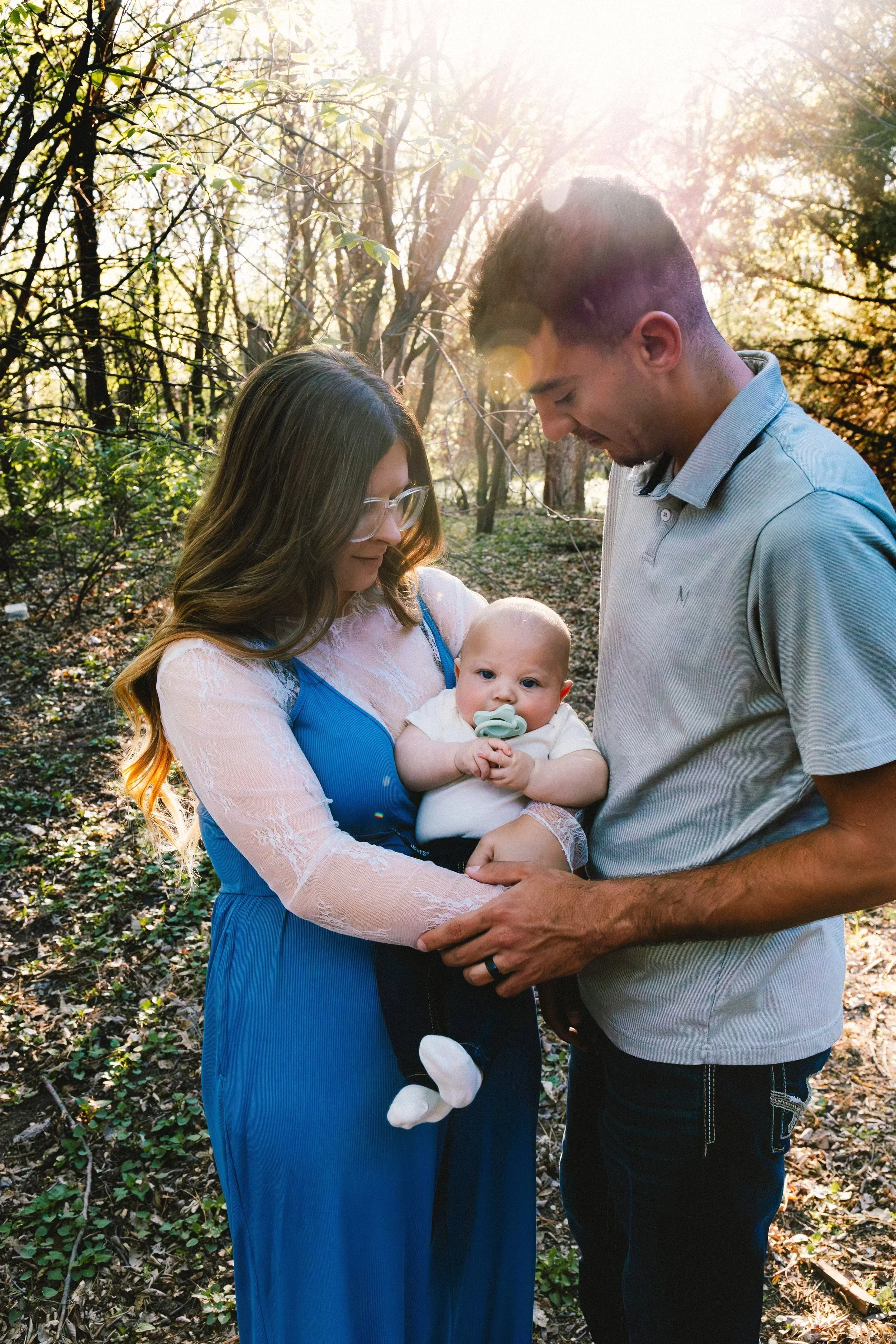 A family of three in a wooded park, with sunlight streaming through the trees. The mother holds a baby with a pacifier, while the father stands close, both looking fondly at the baby.