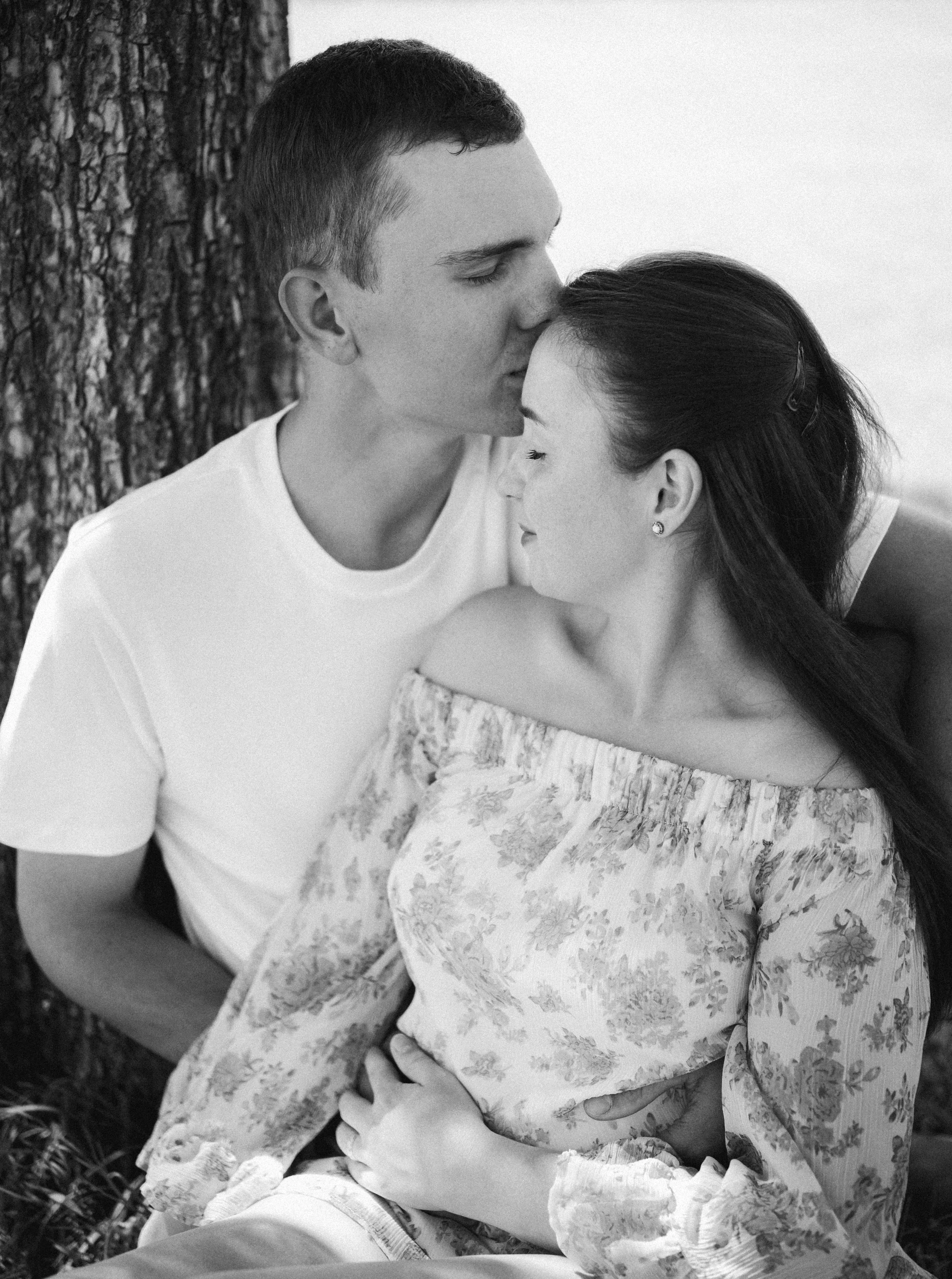 A black and white photo of a man kissing a woman on her forehead. They are sitting close together outdoors near a tree. The woman has long dark hair and is wearing a floral dress, while the man has short hair and is wearing a plain t-shirt.