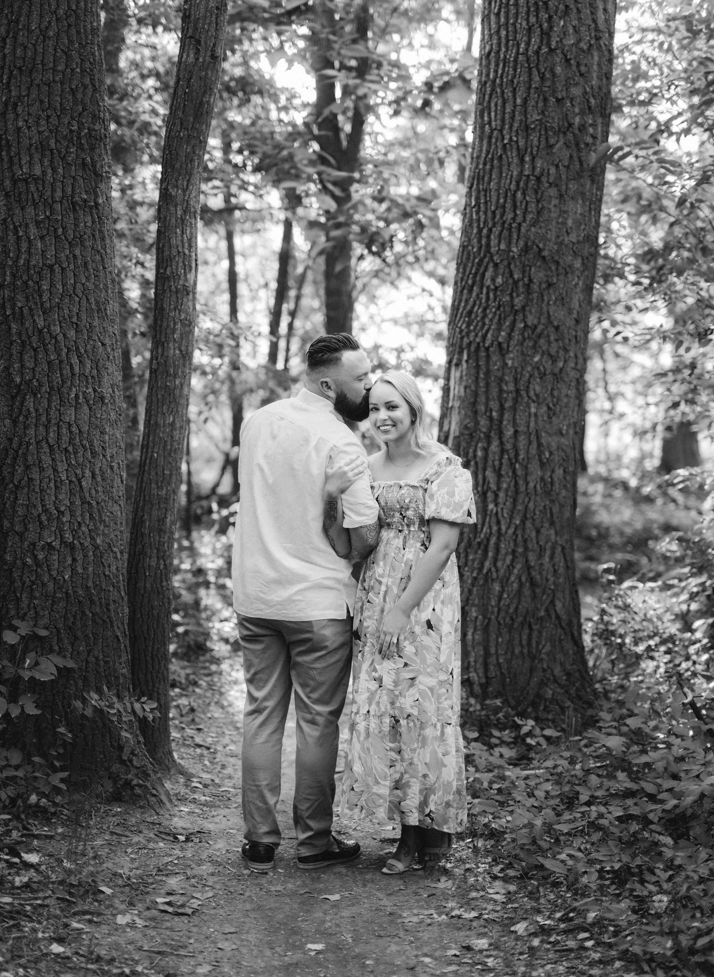 A couple standing on a forest trail, surrounded by tall trees and greenery, sharing a joyful moment with the man kissing the woman's forehead and both smiling.