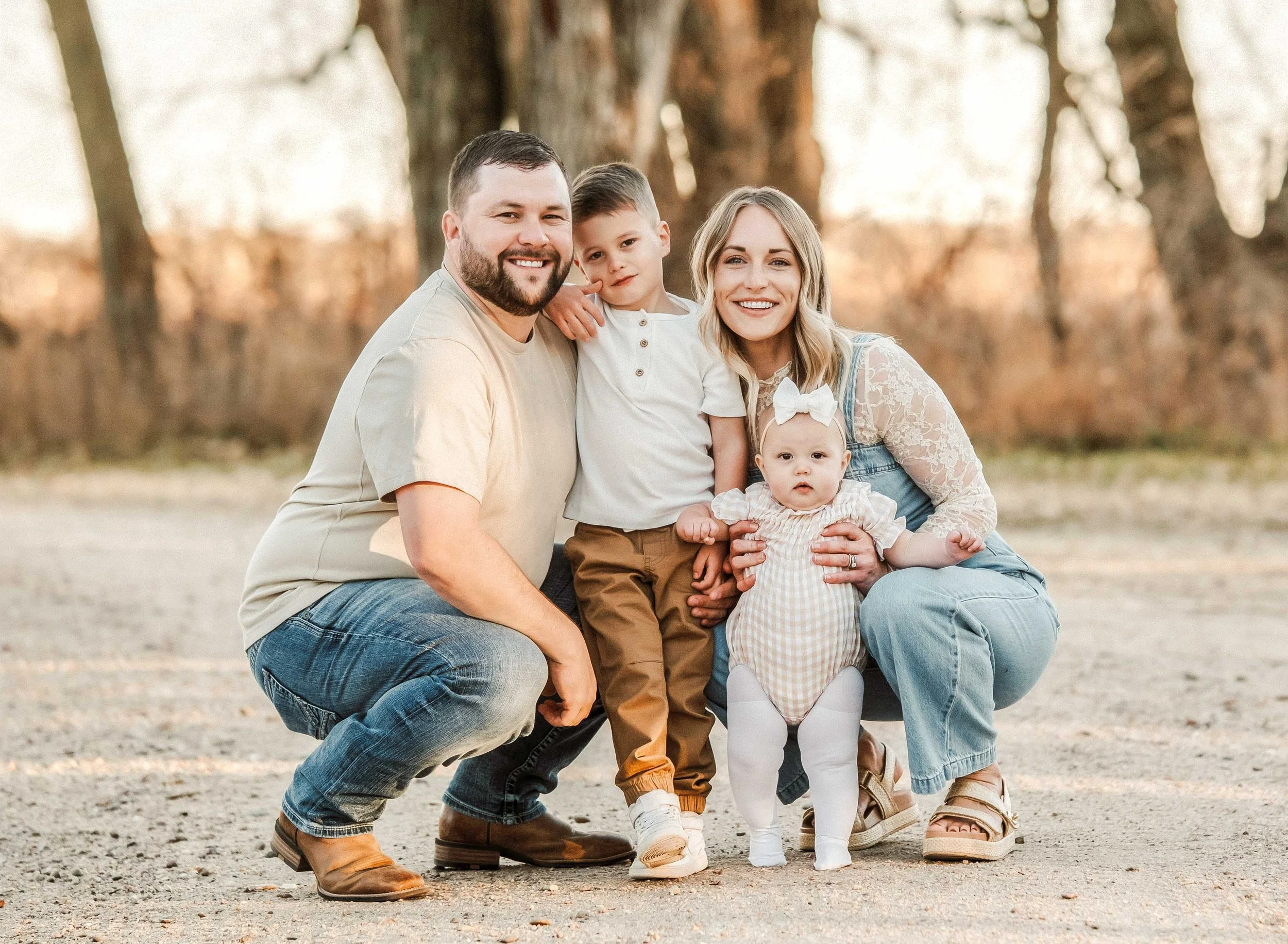 A family of four posing outdoors in a park during fall, with trees in the background. The father has a beard and is wearing a beige t-shirt and jeans. The mother has blonde hair and is dressed in a lace top and denim overalls. Their young son is wear