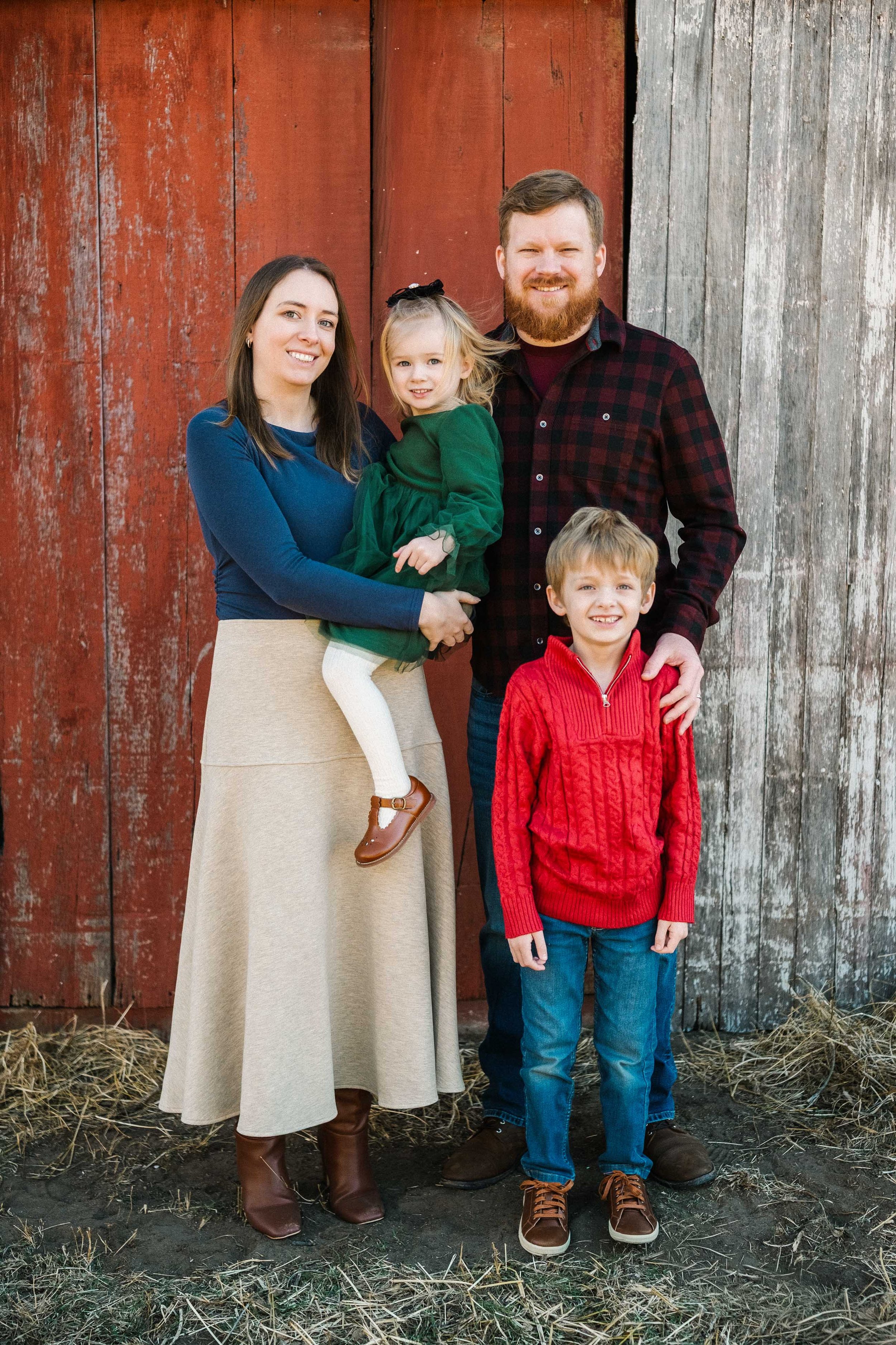 Family of four standing outdoors against a rustic wooden barn, smiling and posing for a photo.