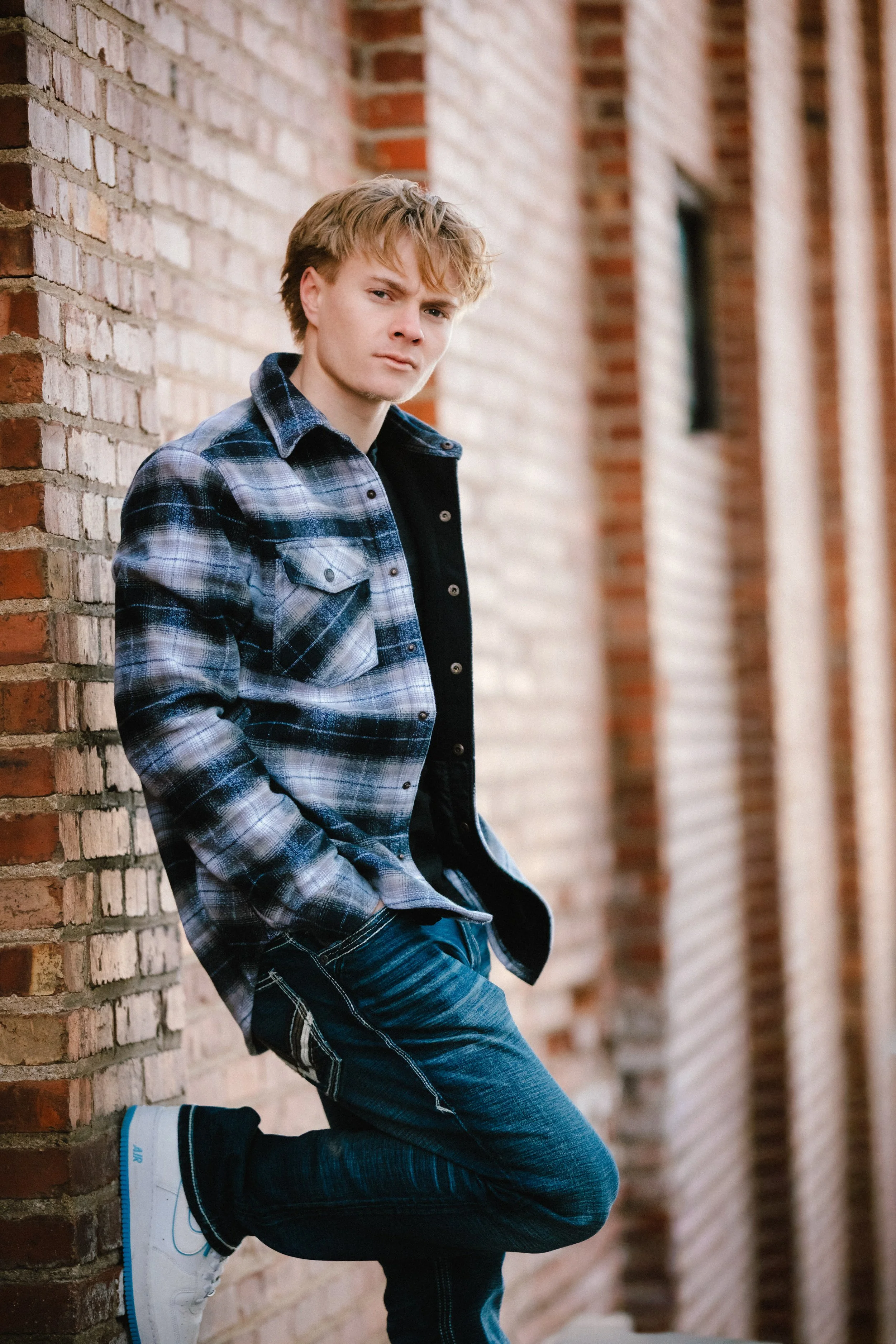 A young man leaning against a brick wall, wearing a blue plaid shirt, jeans, and white sneakers, looking at the camera with a serious expression.