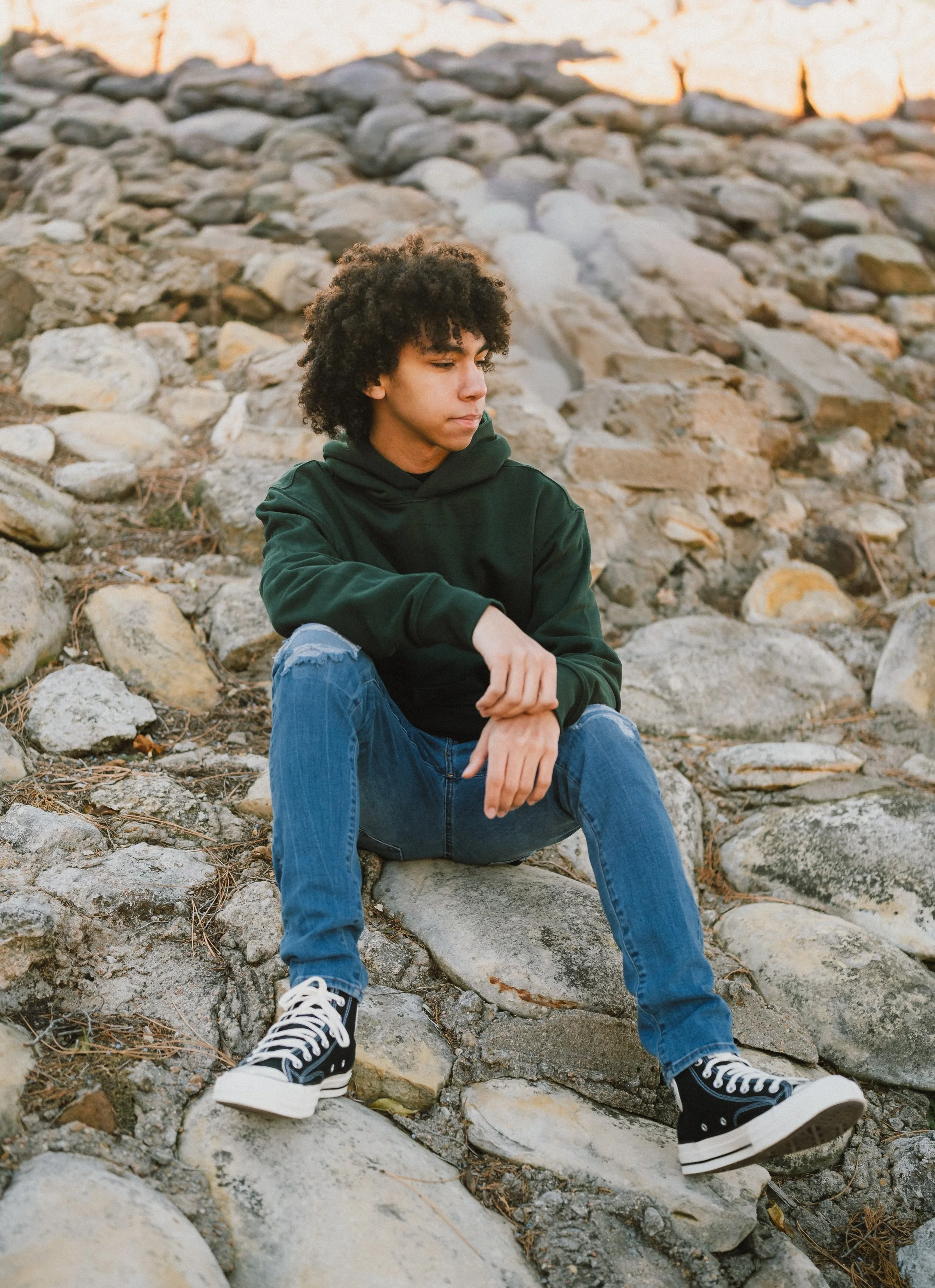 A young person with curly hair sitting on rocks near the water, wearing a green hoodie, ripped jeans, and black and white sneakers.