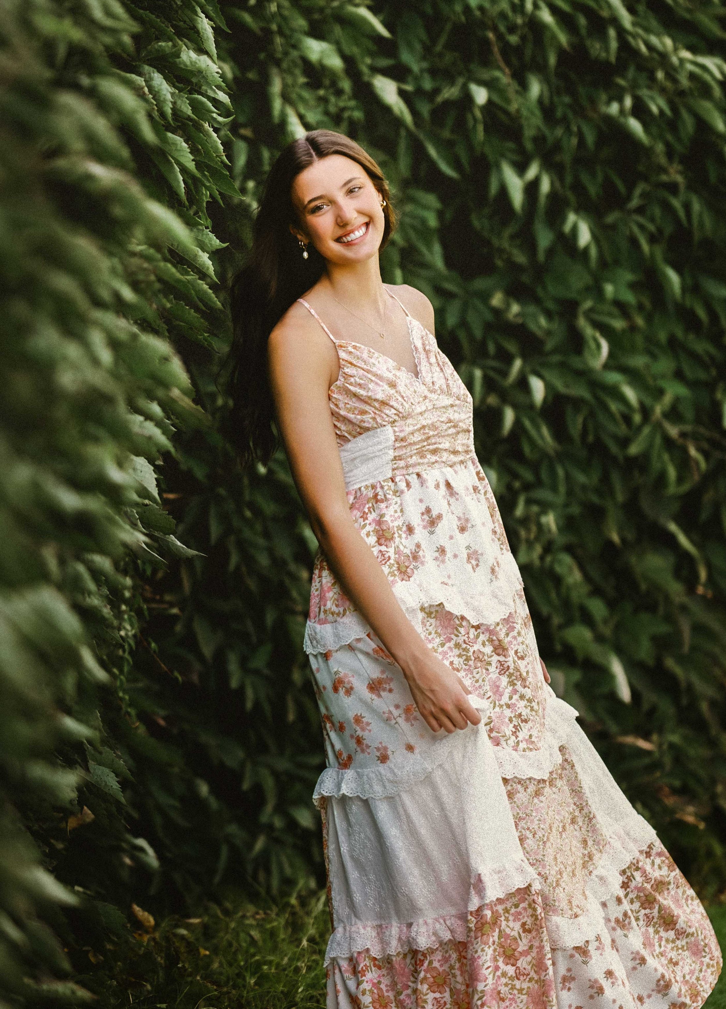 A young woman in a layered floral dress with spaghetti straps, smiling and leaning against a hedge in a lush outdoor setting.