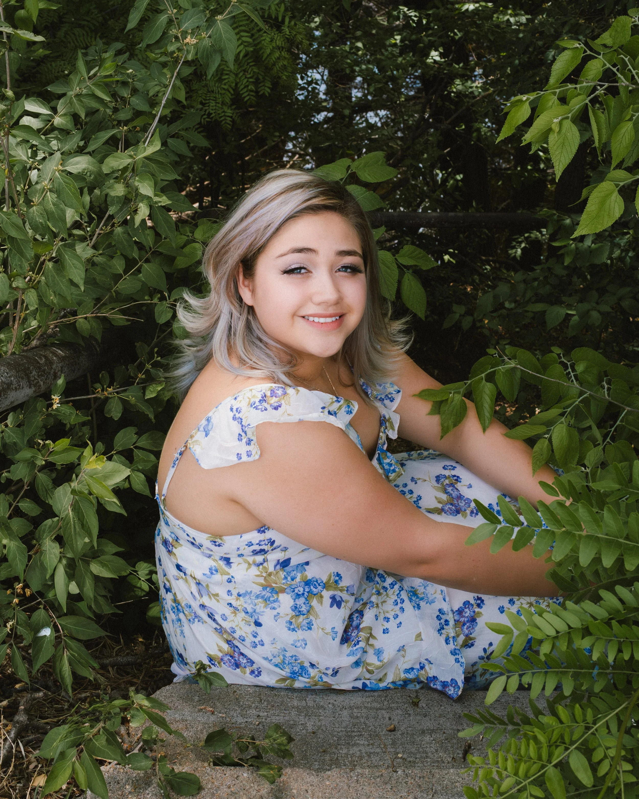 A woman with blonde hair smiling and looking at the camera while sitting outdoors among green foliage, wearing a white dress with blue and yellow floral pattern.