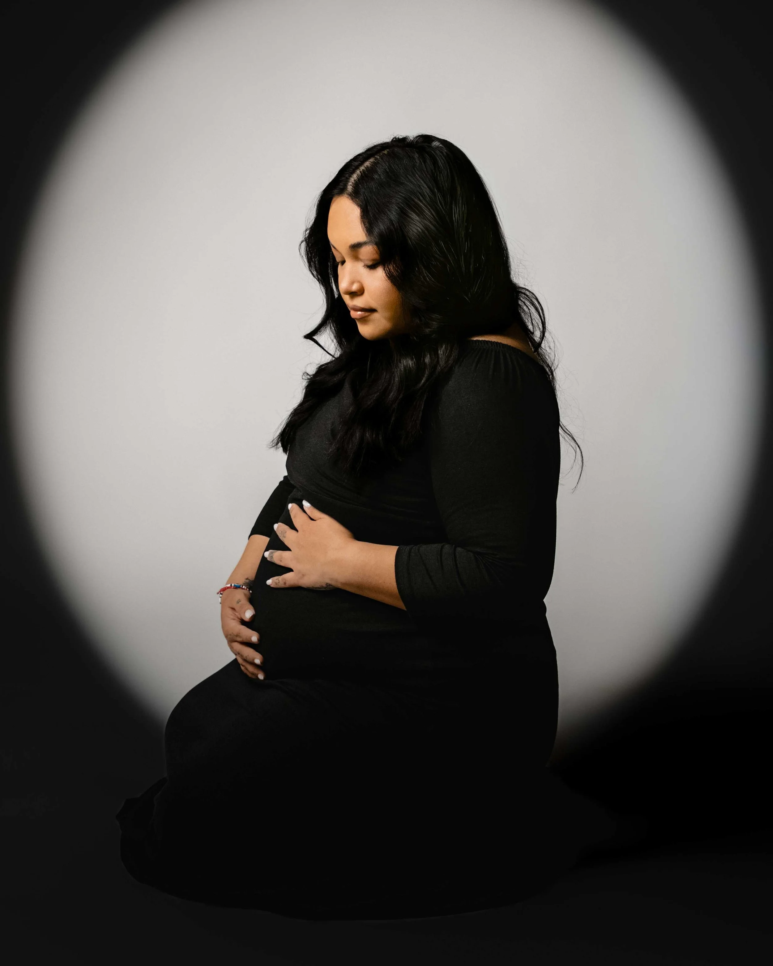 A pregnant woman with long black hair and dark clothing, sitting on her knees with her hands resting on her belly, looking down with a serene expression.