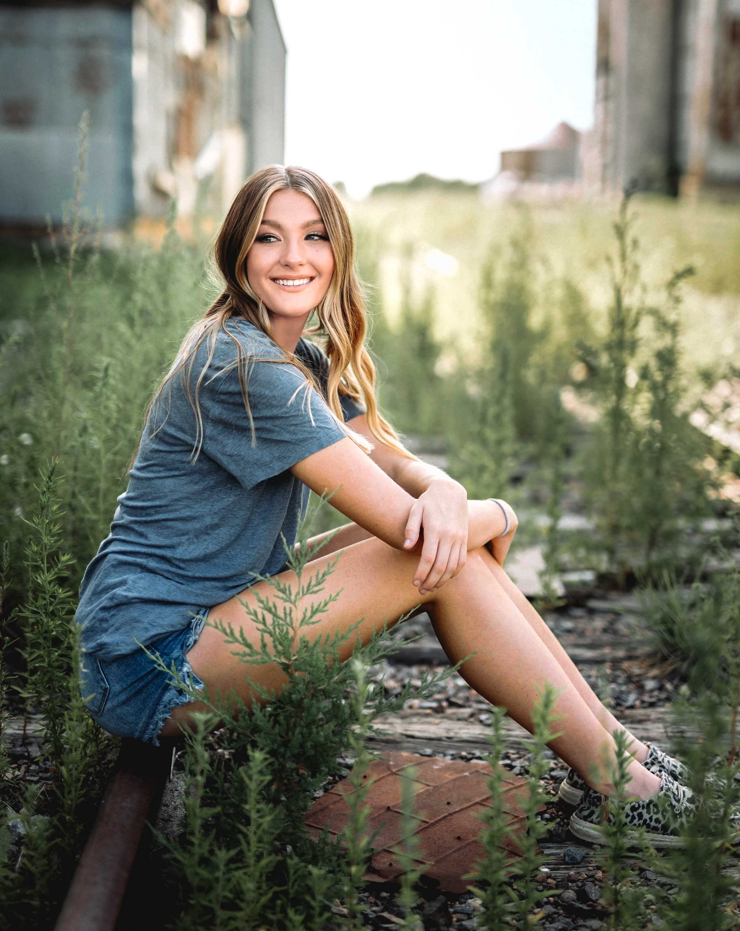 A young woman with long blonde hair, wearing a blue t-shirt, denim shorts, and sneakers, sitting on the ground among green plants on an abandoned railway track with old buildings in the background, smiling and looking away.