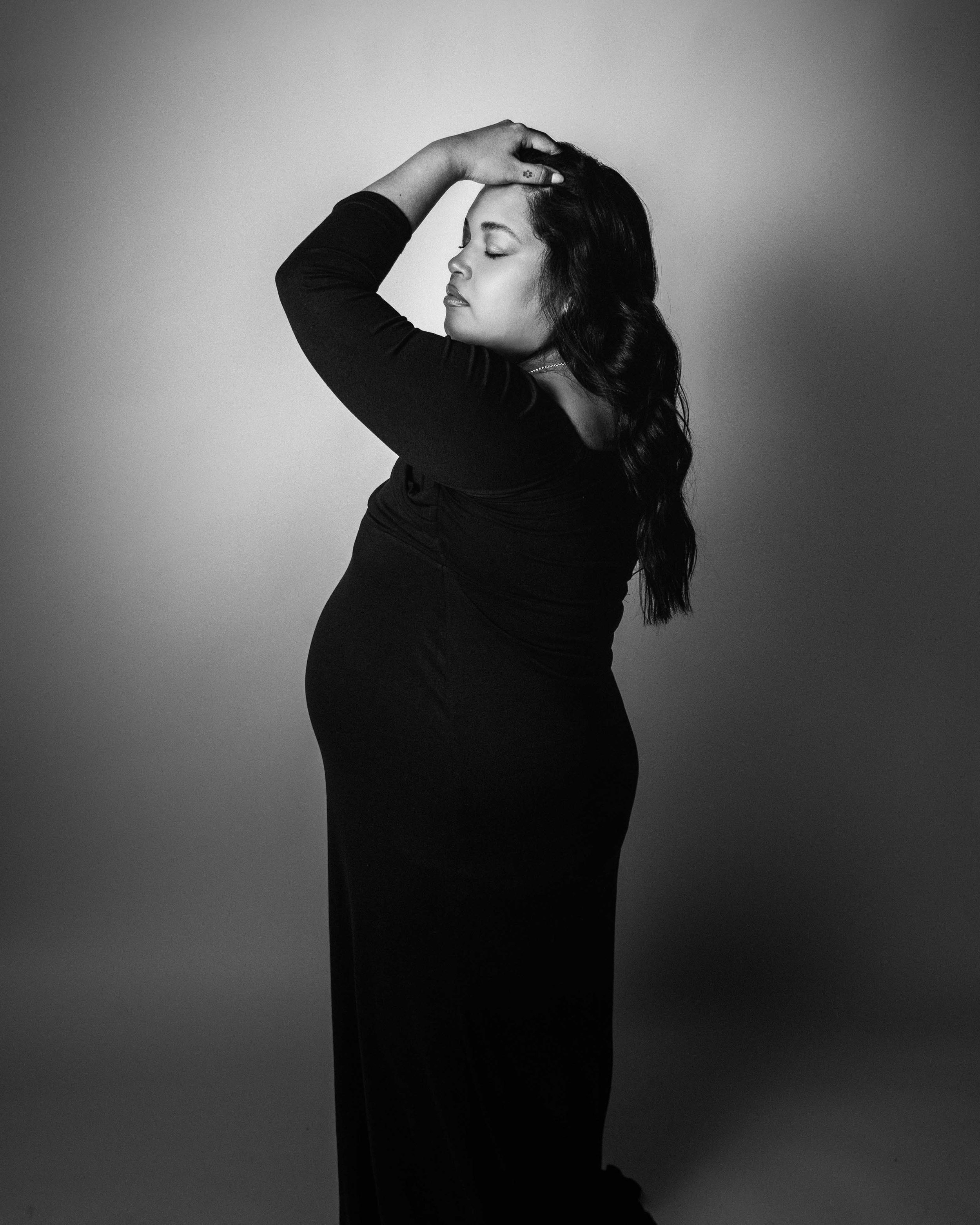 A woman with long dark hair posing with one hand on her head and eyes closed, dressed in black, in a black and white studio portrait.