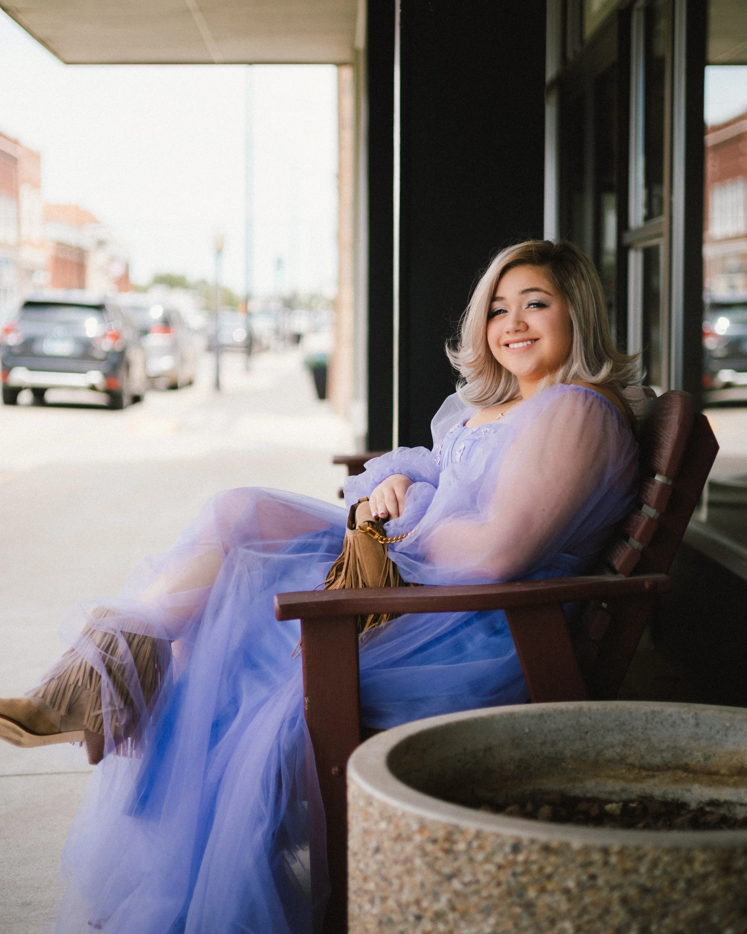 Young woman with blonde hair sitting on porch, smiling and wearing a lavender dress, with a brown fringed purse on her lap, next to a table with a stone mortar and pestle, outside a building with cars parked on the street.