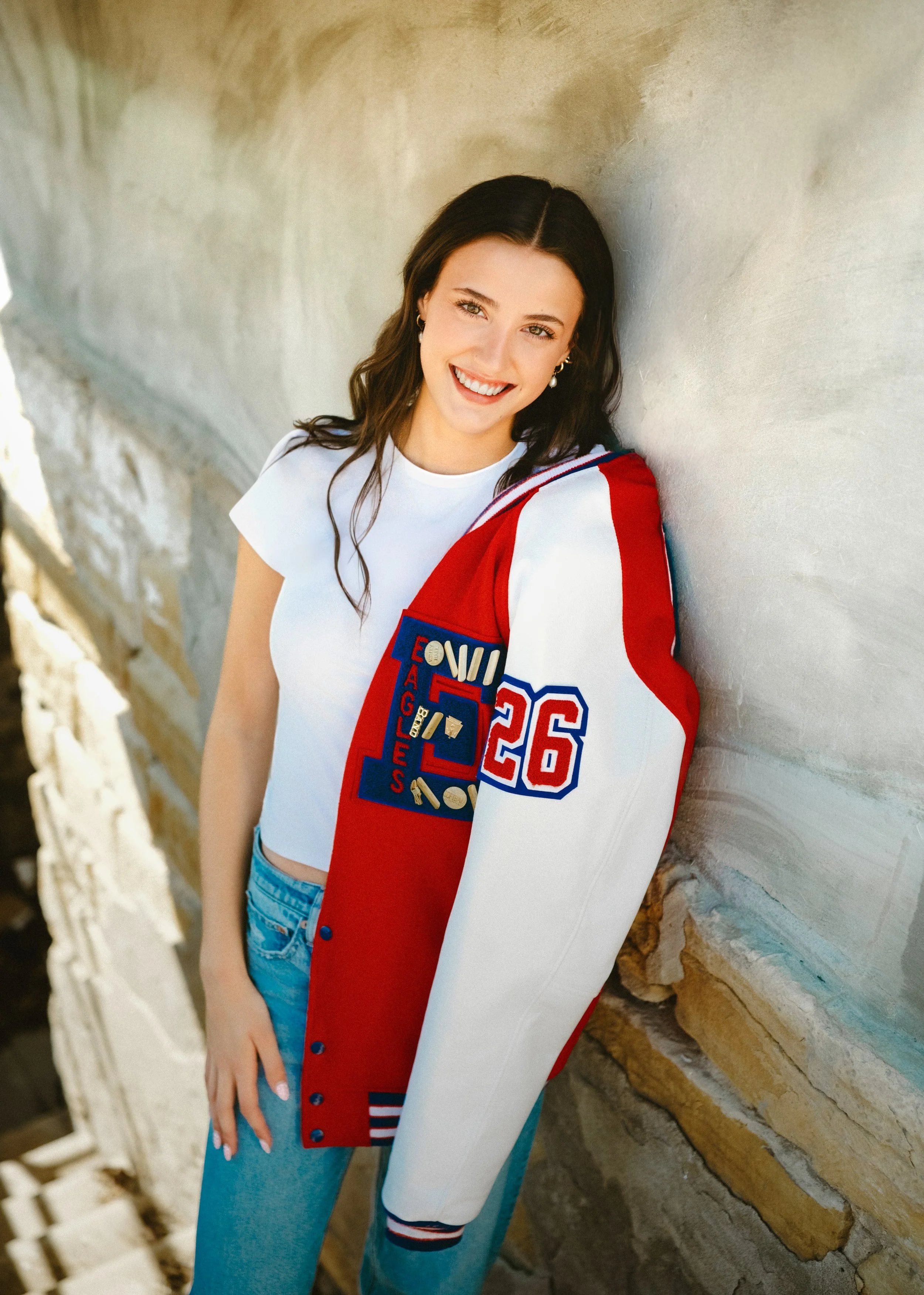Young woman with dark hair and a white T-shirt smiling while leaning against a textured beige wall, holding a red varsity jacket with blue and white accents and the number 26.
