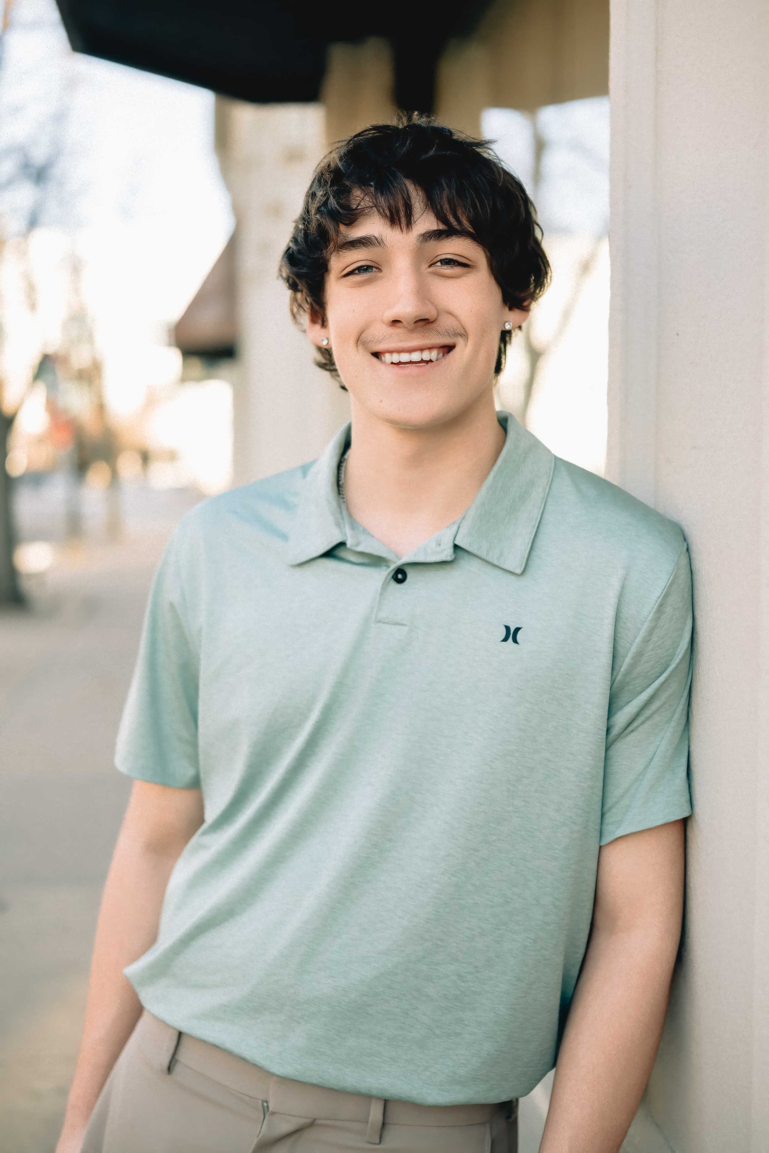 Young man with dark, wavy hair, wearing a light green polo shirt and beige pants, smiling and leaning against a wall outdoors.