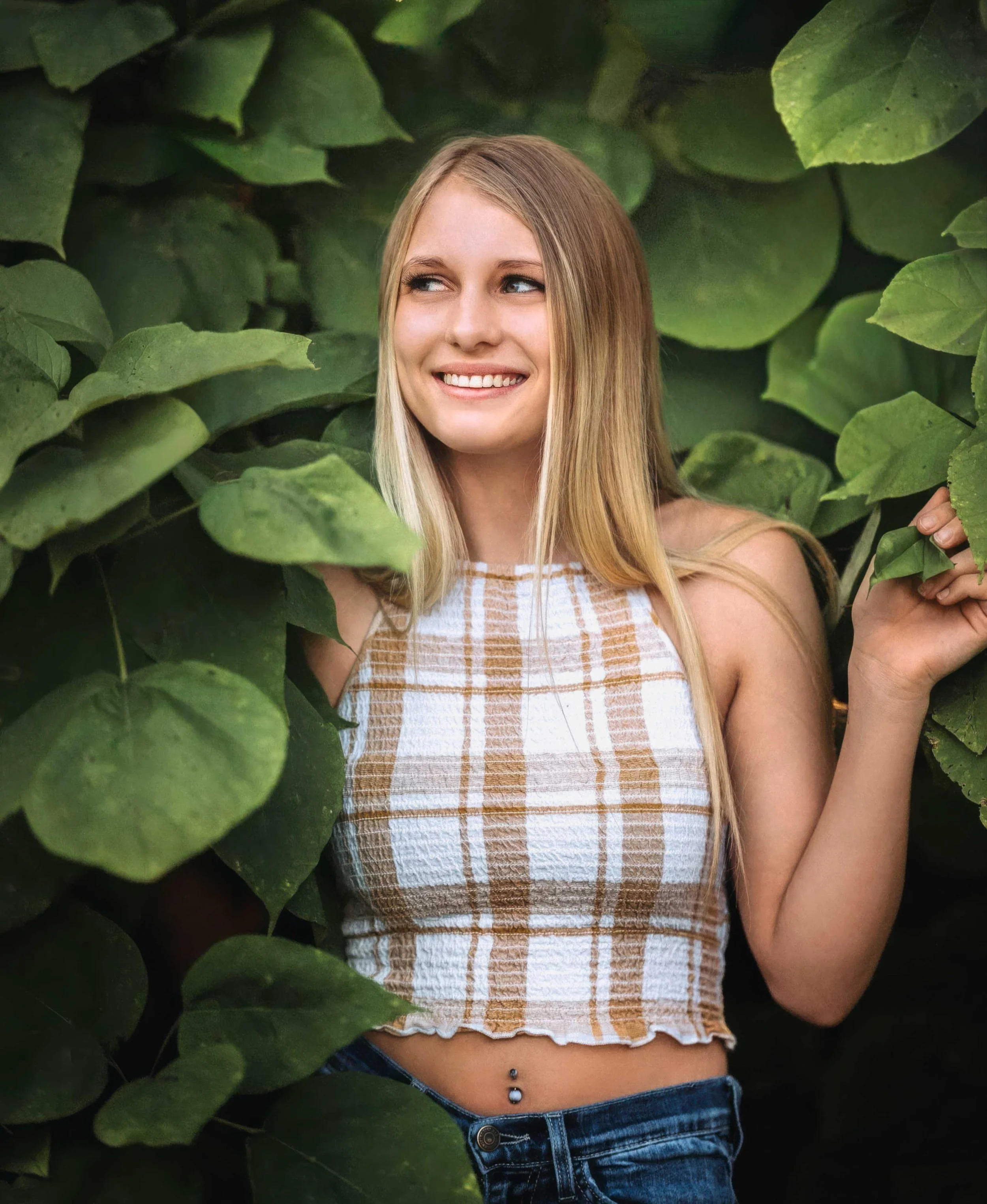 A young woman with long blonde hair smiling among green leafy plants, wearing a sleeveless plaid top and jeans with a belly piercing.