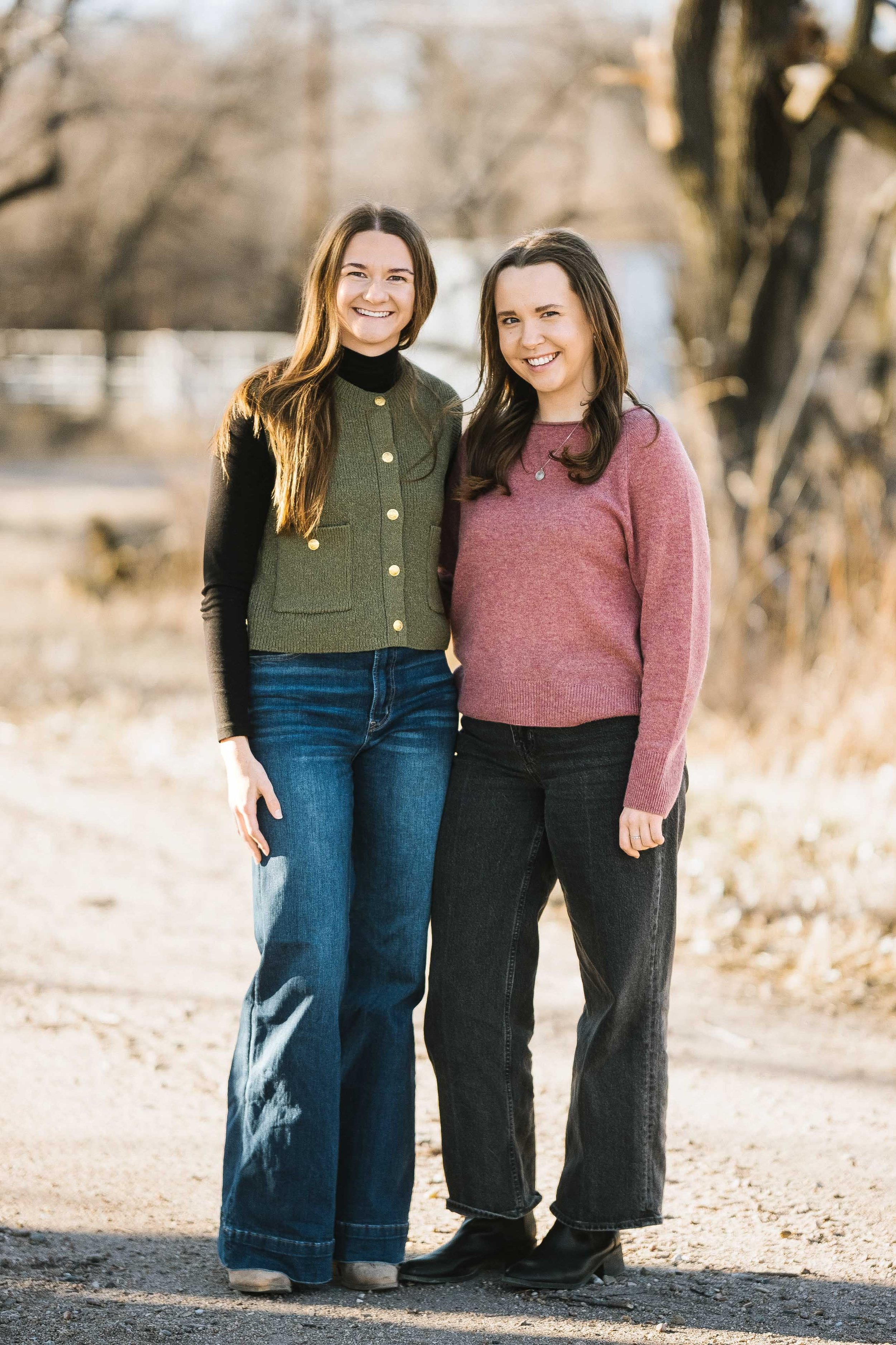 Two women standing outdoors on a dirt path, smiling, with trees in the background. one wearing a patterned colorblock cardigan and jeans, the other in a pink sweater and black jeans.