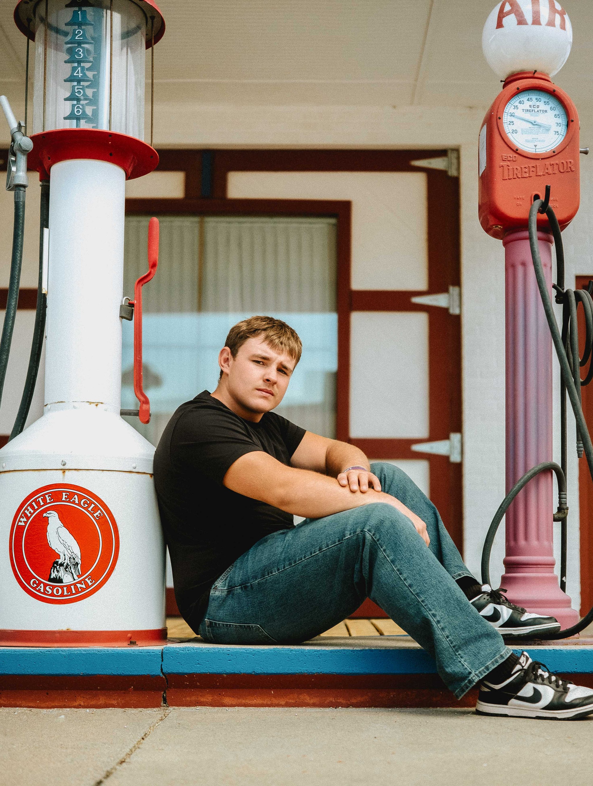 A young man sitting on the ground next to a vintage White Eagle Gasoline pump at a gas station, with a serious expression on his face.