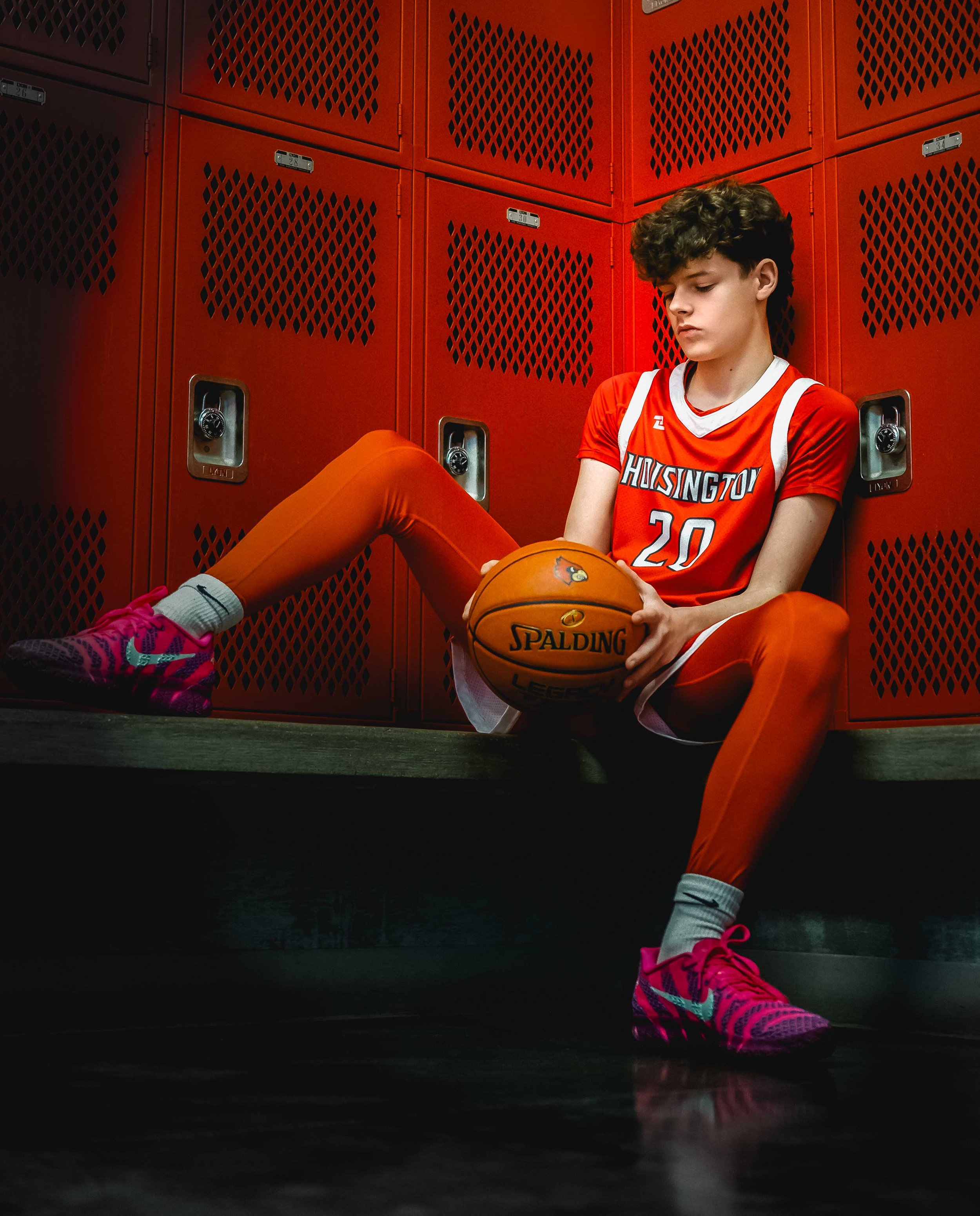 A teenage basketball player wearing a red and white uniform sitting on the floor of a locker room with red lockers behind him, holding a basketball.