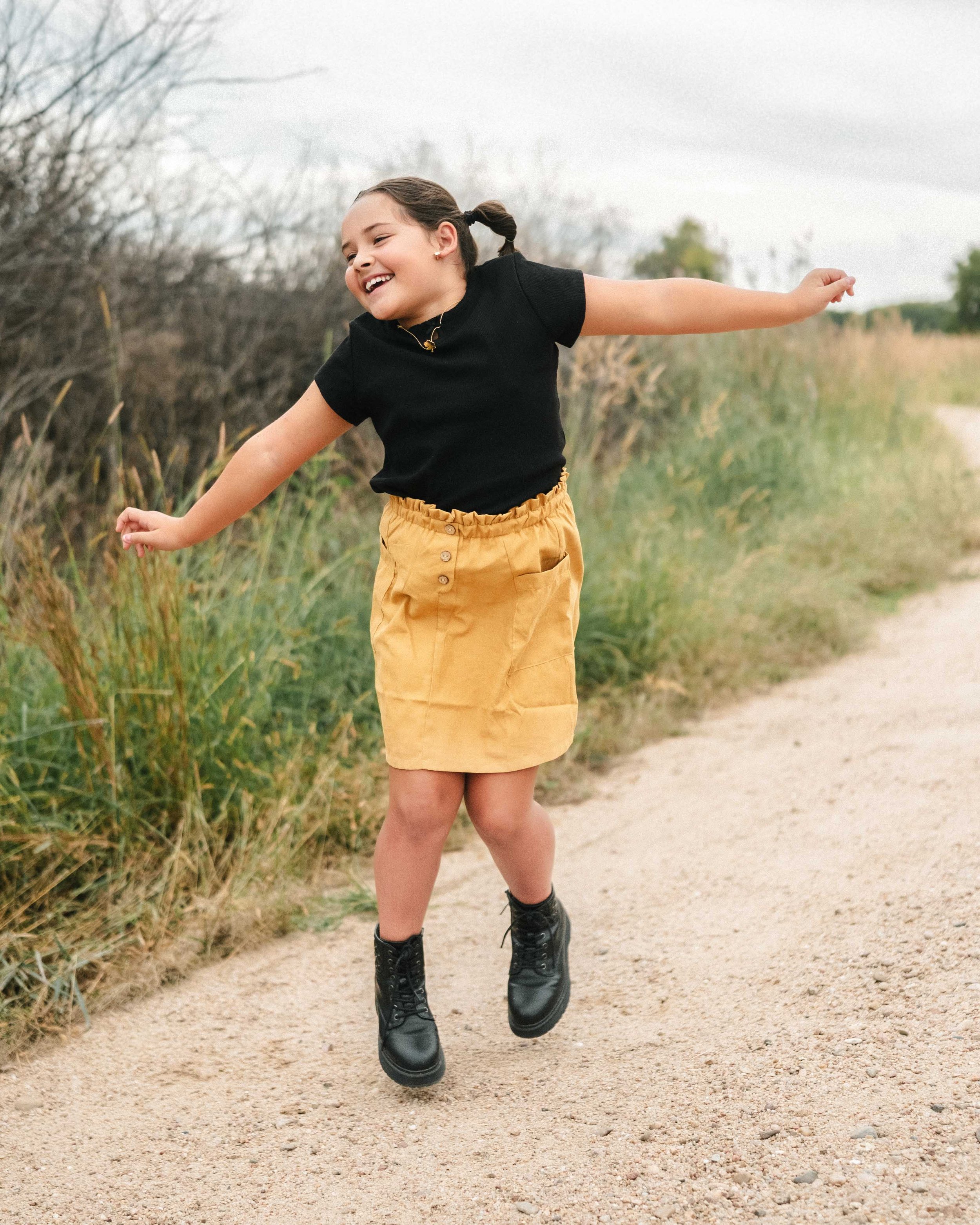 Young girl joyfully jumping on a dirt path with greenery and bushes on either side under an cloudy sky.