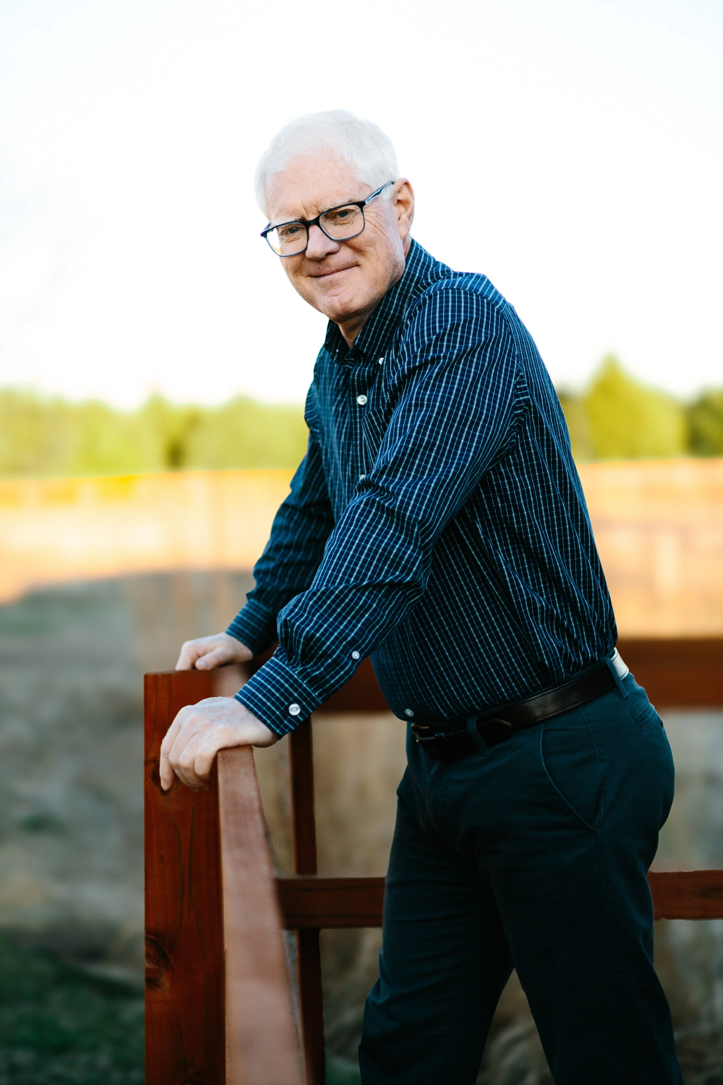 Older man with white hair and glasses leaning on a wooden railing outdoors.