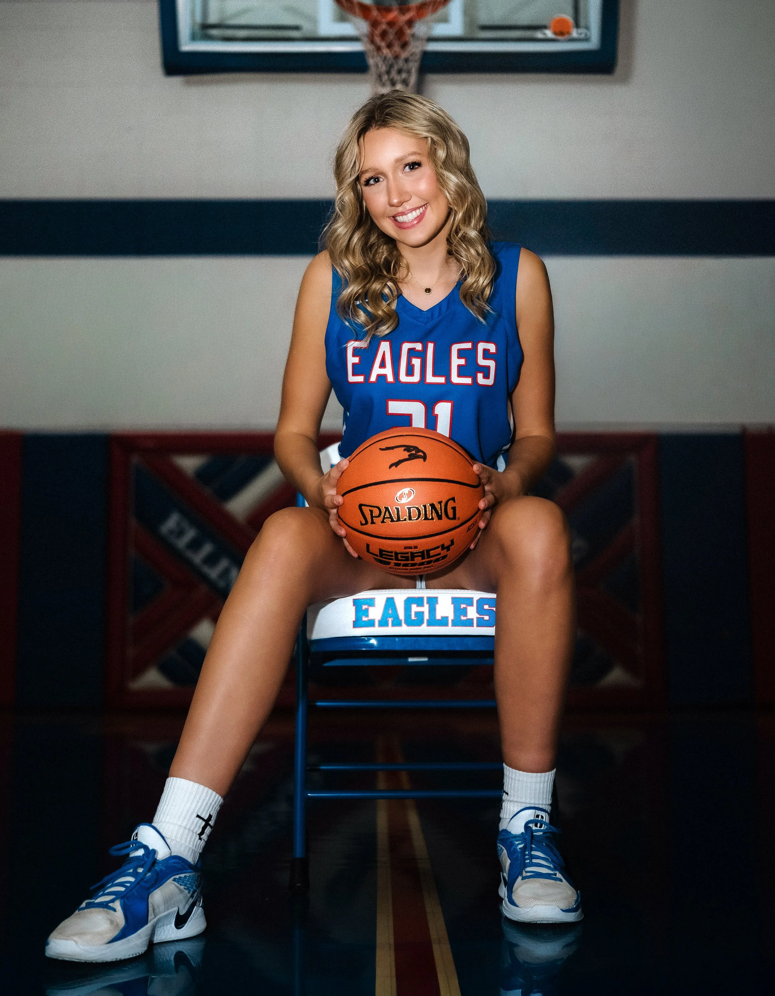 A young woman with blonde curly hair in a blue basketball jersey, sitting on a chair on a basketball court, holding a basketball, smiling.