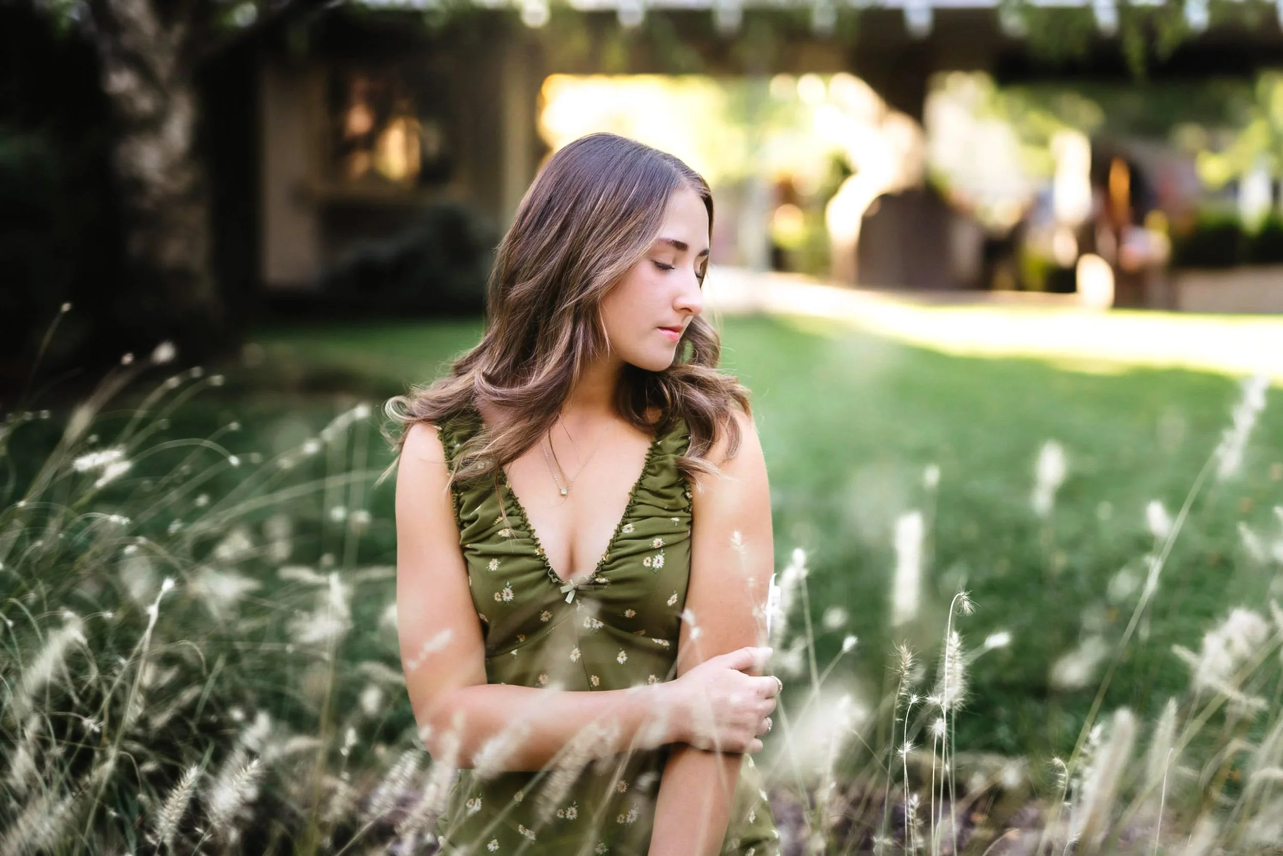 A young woman with wavy brown hair in a green dress standing outdoors in a grassy area with tall, white, fluffy plants, with a blurred background of trees and a house.
