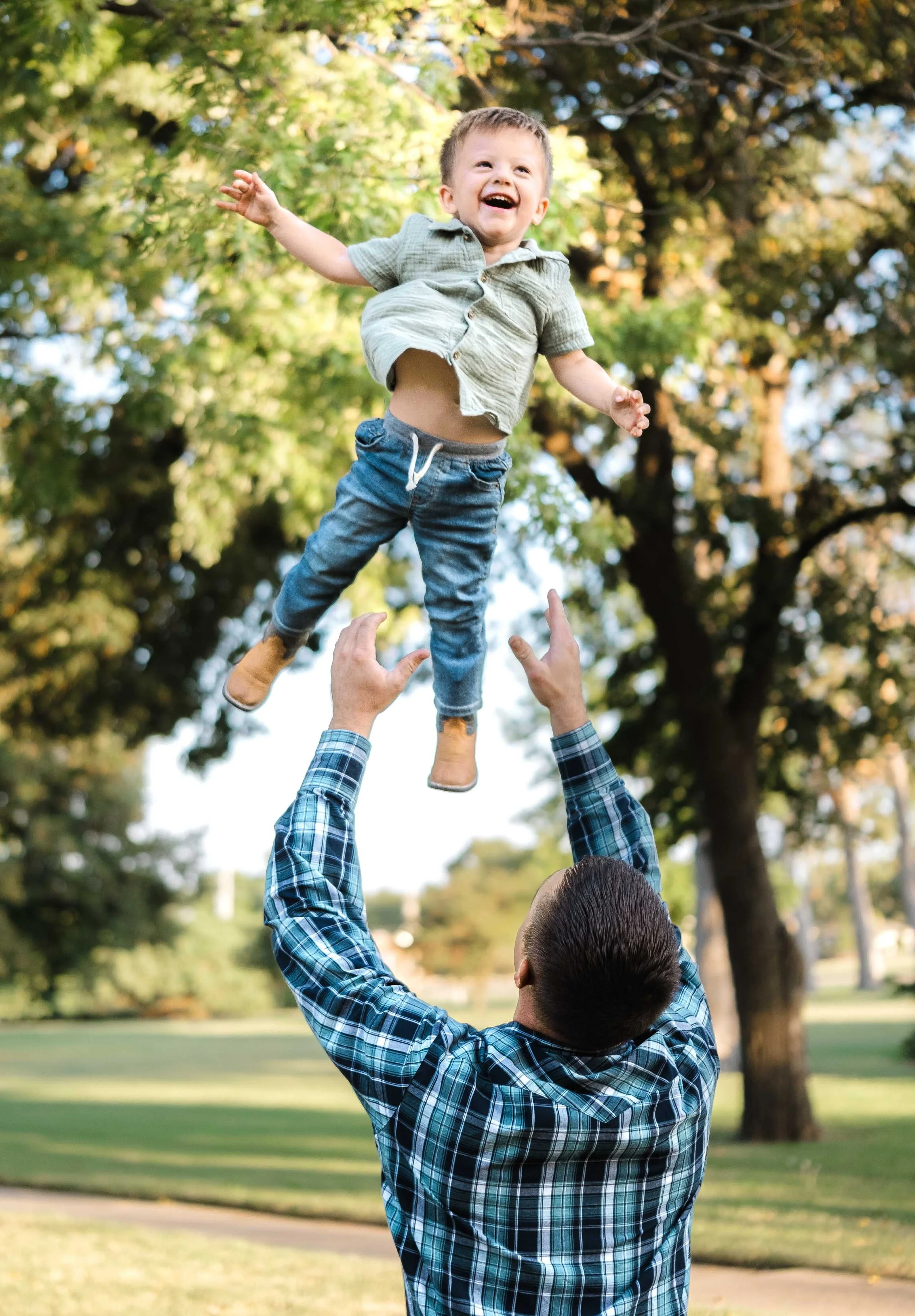 A man is tossing a young boy into the air outdoors in a park surrounded by trees, with both smiling and appearing joyful.