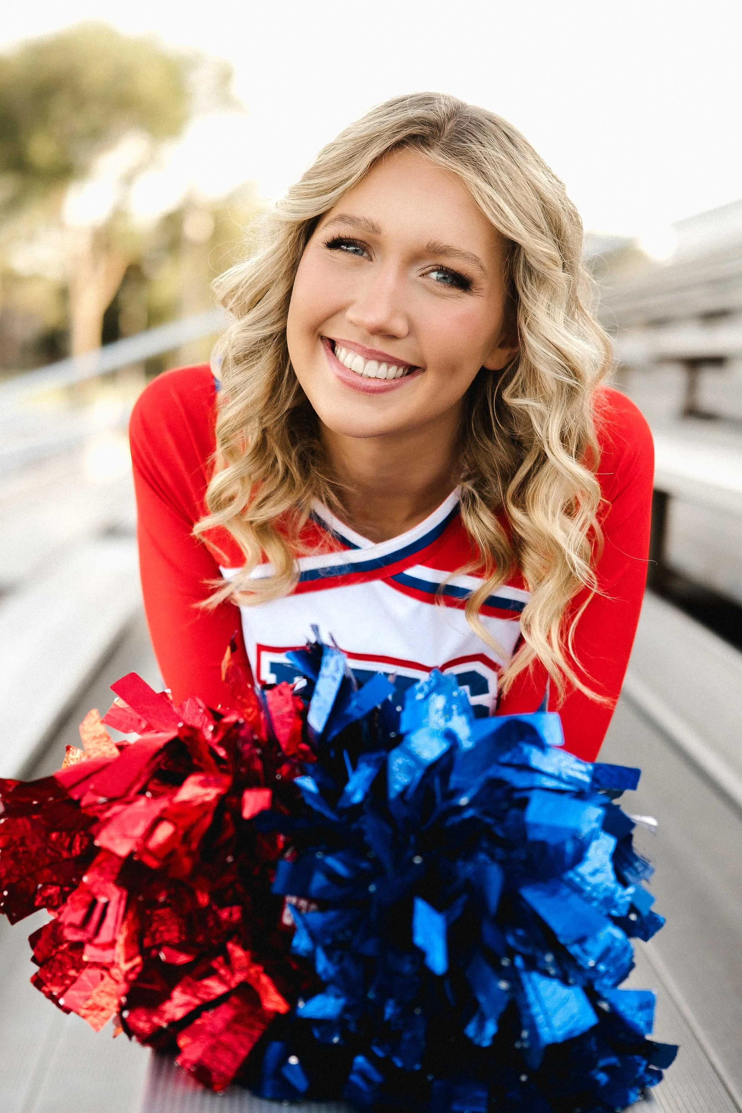 A smiling young woman in a cheerleading uniform and pom-poms sitting on bleachers.