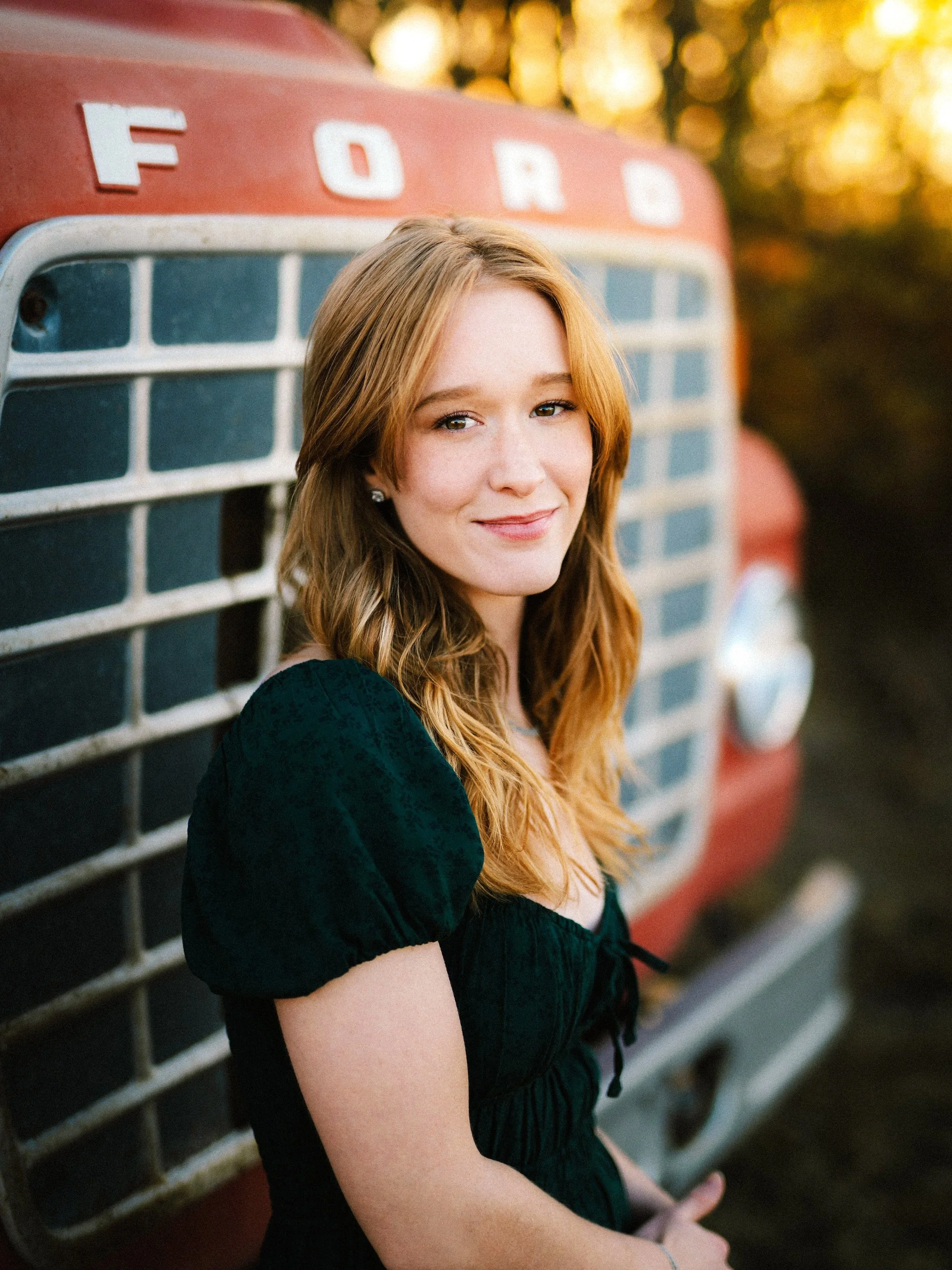 A young woman with long, wavy red hair and fair skin smiling while sitting in front of a vintage red Ford truck outdoors during sunset.
