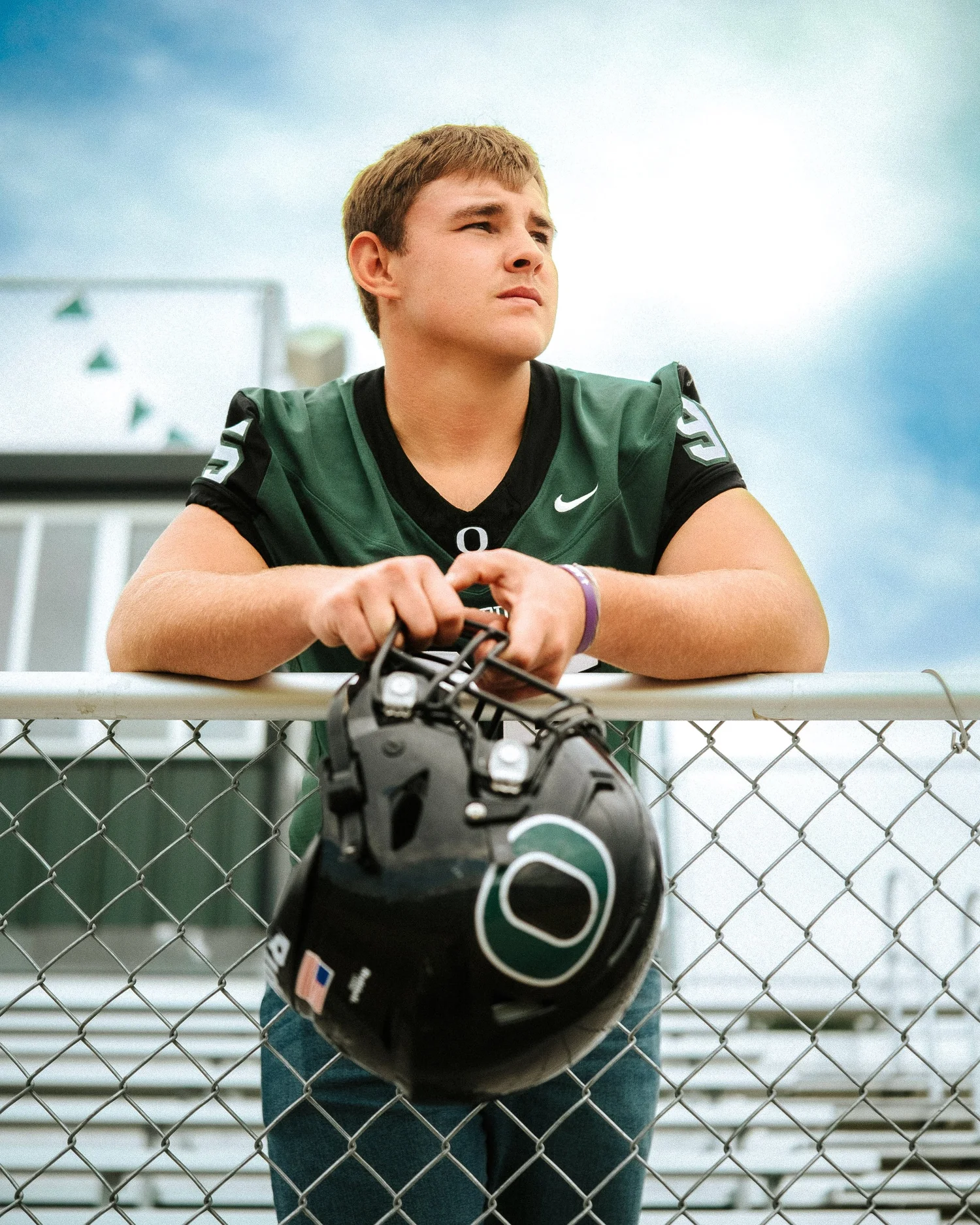 A senior football player leaning on a chain-link fence holding his helmet