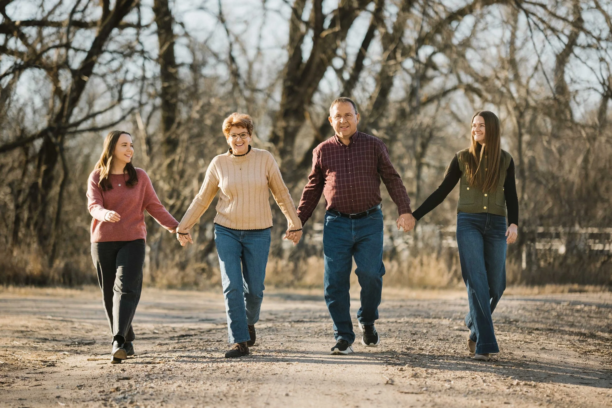 A family of four walking outdoors on a dirt path during daytime. The family includes two young women, a middle-aged woman, and a middle-aged man. They are holding hands and smiling, surrounded by leafless trees in the background.