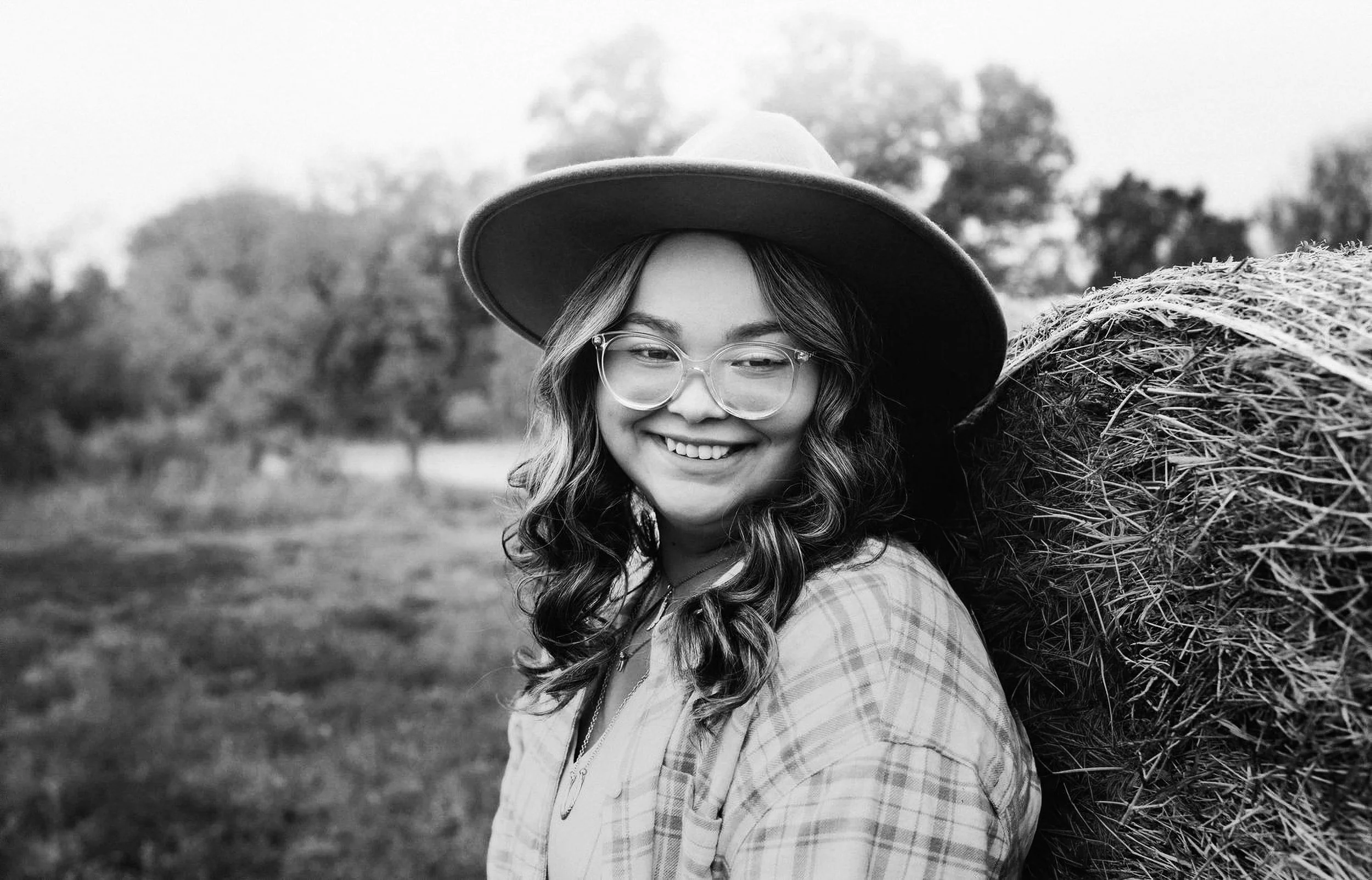 Black and white photo of a smiling woman with glasses and wavy hair, wearing a wide-brimmed hat and a plaid shirt, standing outdoors with a large hay bale on her shoulder and trees in the background.