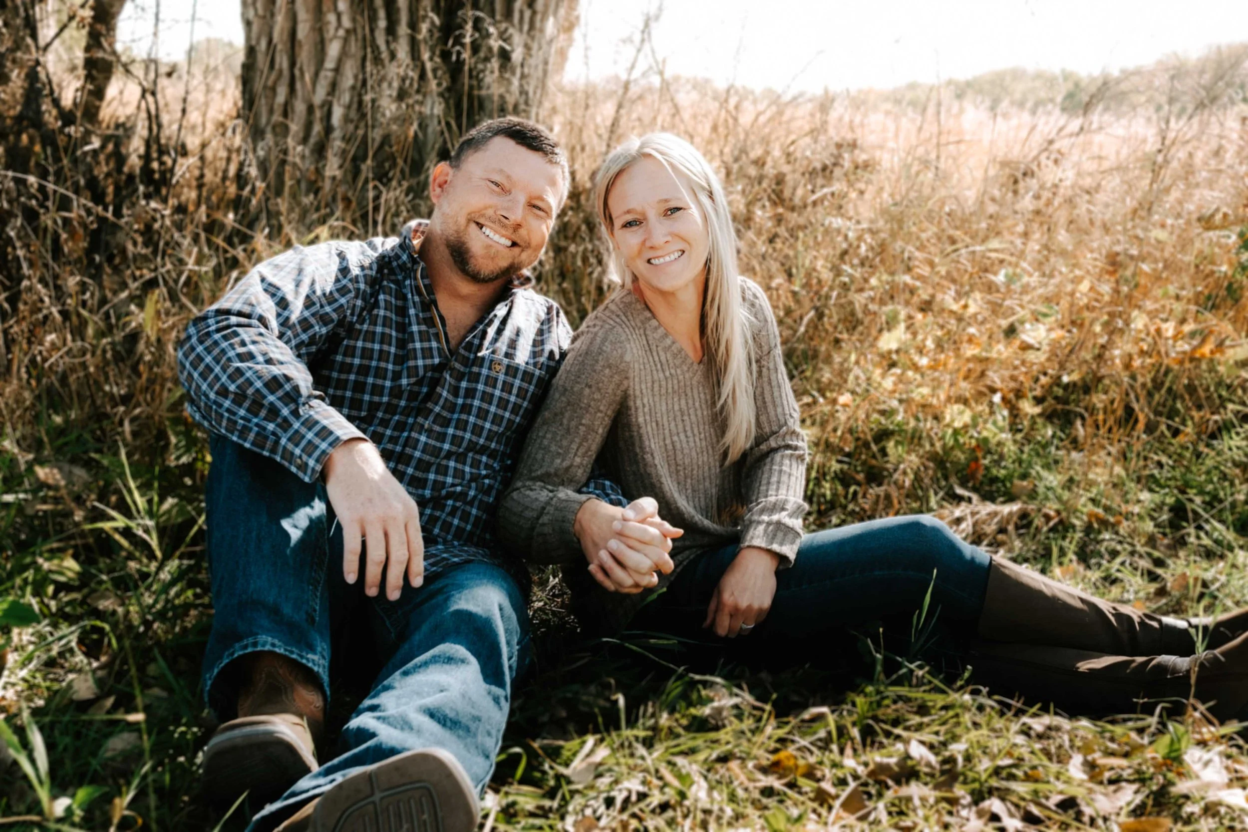 A smiling man and woman sitting outdoors in a grassy area with tall dry grass and trees in the background, holding hands and looking at the camera on a sunny day.