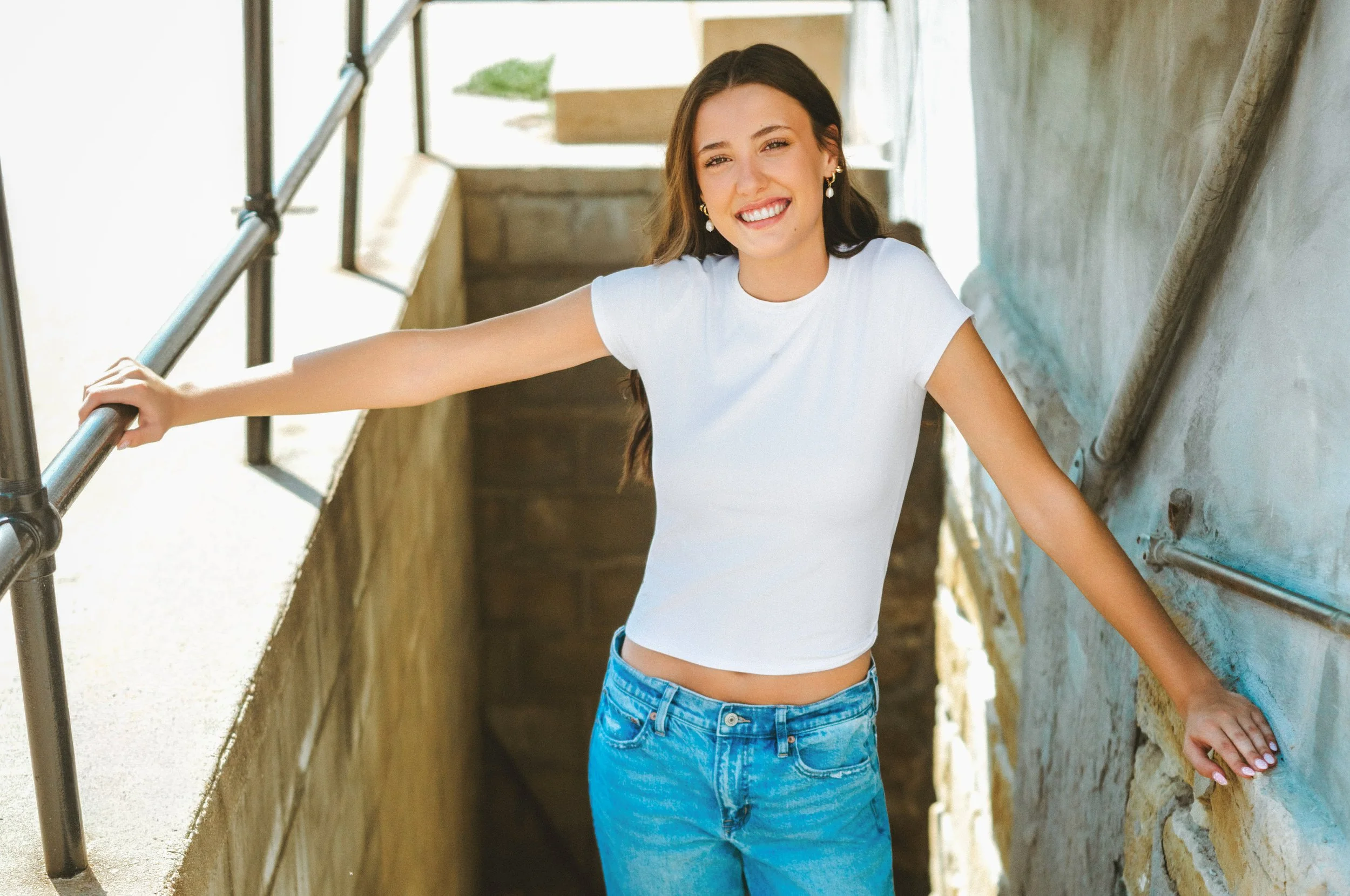 A young woman with long brown hair in a white t-shirt and blue jeans smiling while holding onto a metal railing in an outdoor stairwell.