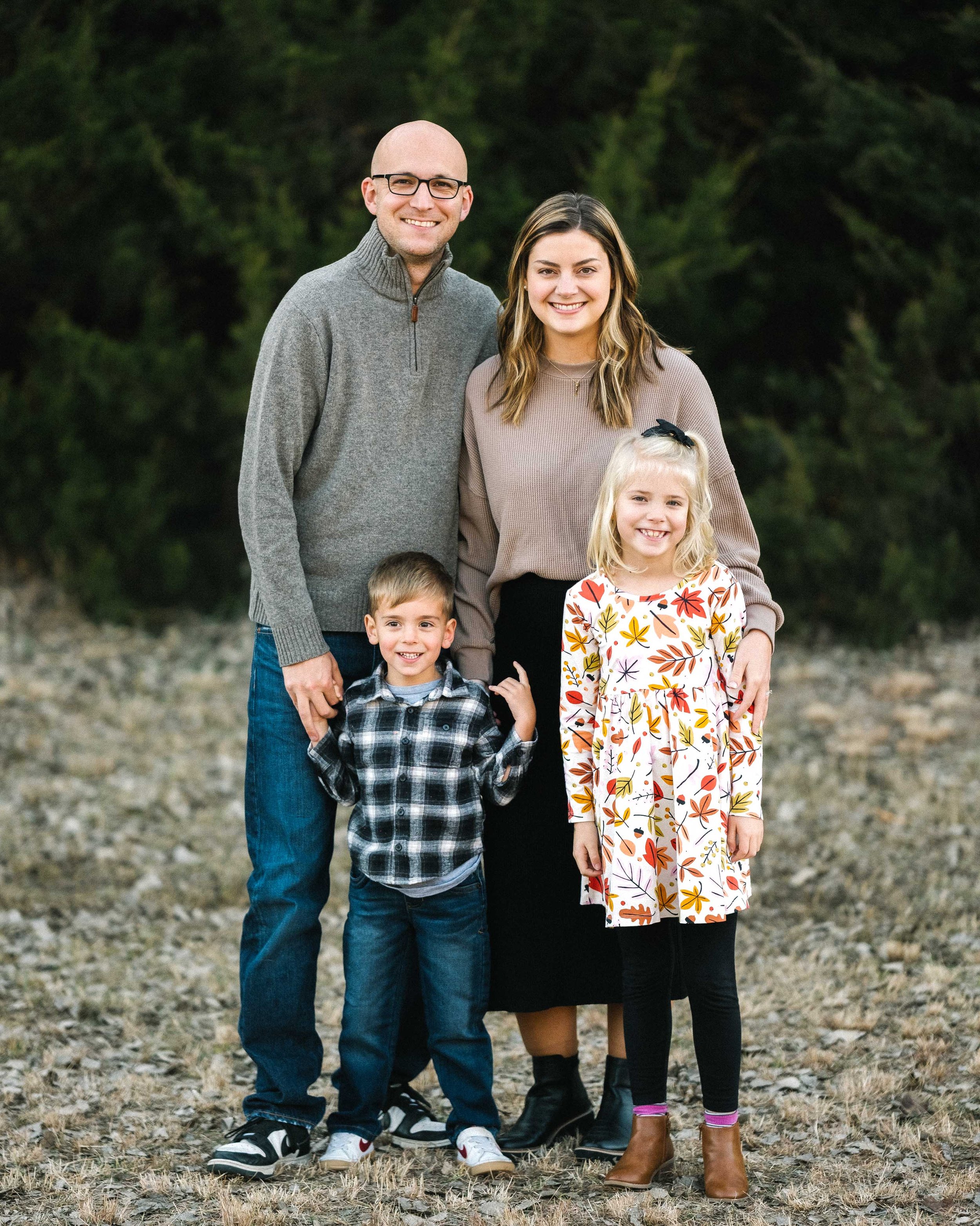 A family of five standing outdoors on a grassy and rocky area with green trees in the background. The family consists of a man with glasses, a woman, a young boy, and a young girl, all smiling at the camera.