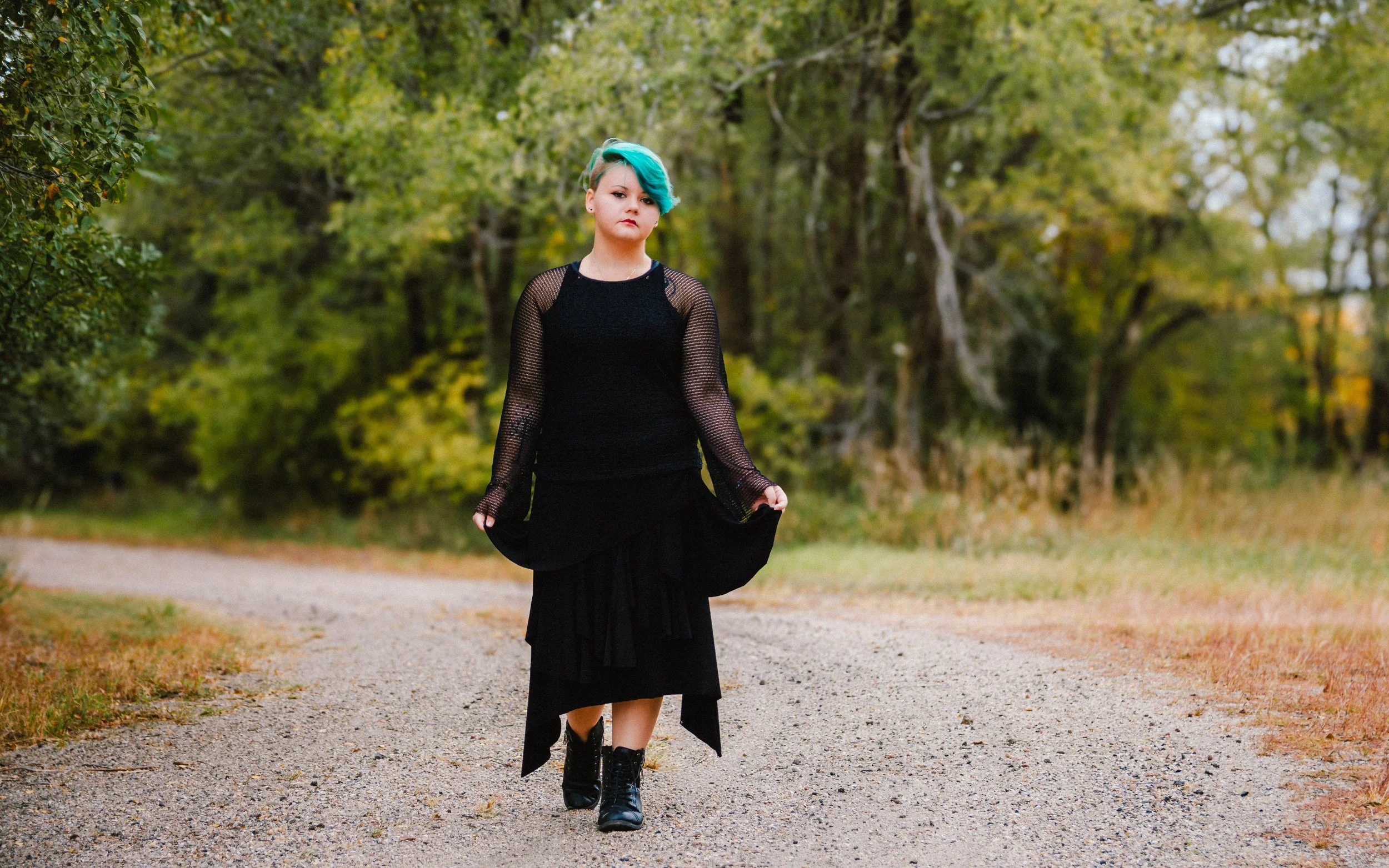 A young woman with teal hair and a black outfit walking on a gravel path surrounded by green trees.