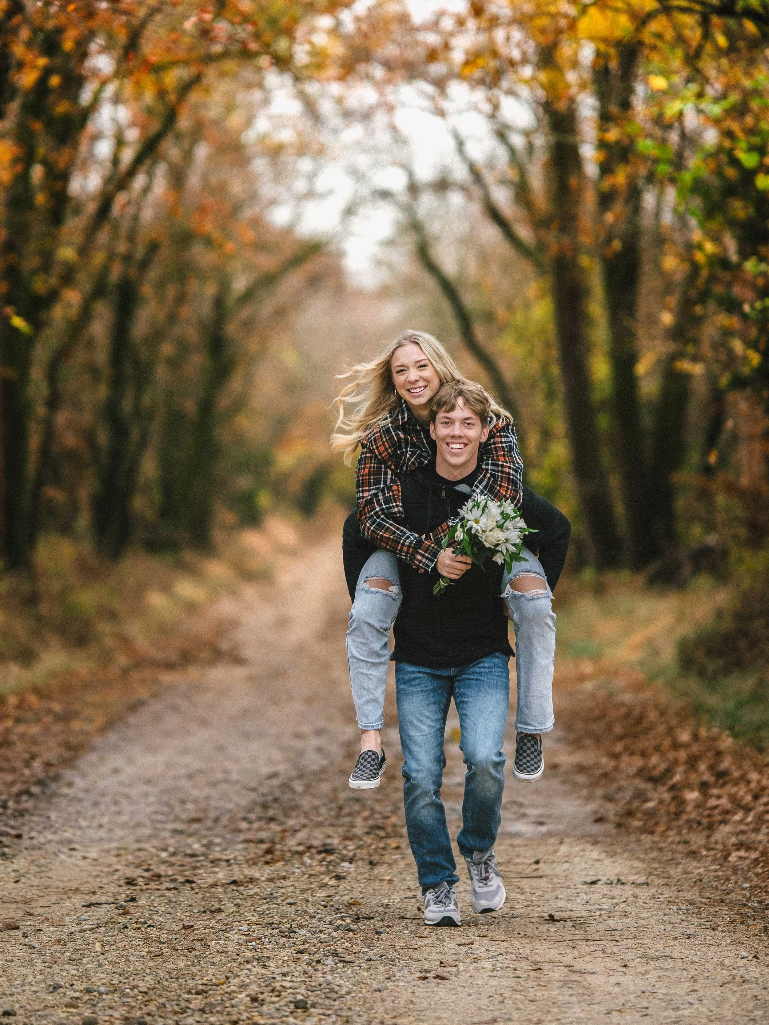 A young couple is enjoying a walk on a forest path during fall, with the woman sitting on the man's back, holding a bouquet of flowers, both smiling and happy.