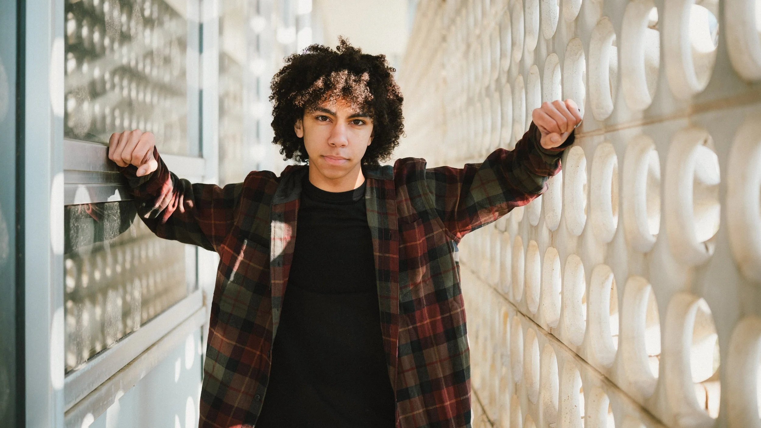 Young person with curly hair standing between glass and concrete walls, looking confidently at the camera with arms raised and hands gripping the walls.