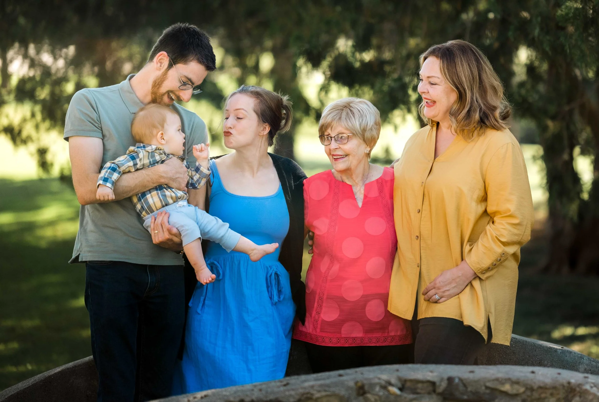 Family gathering outdoors with five members, including a young man holding a baby boy, a young woman in a blue dress, an elderly woman in a red shirt, and a woman in a yellow blouse, standing on a park lawn with trees in the background.