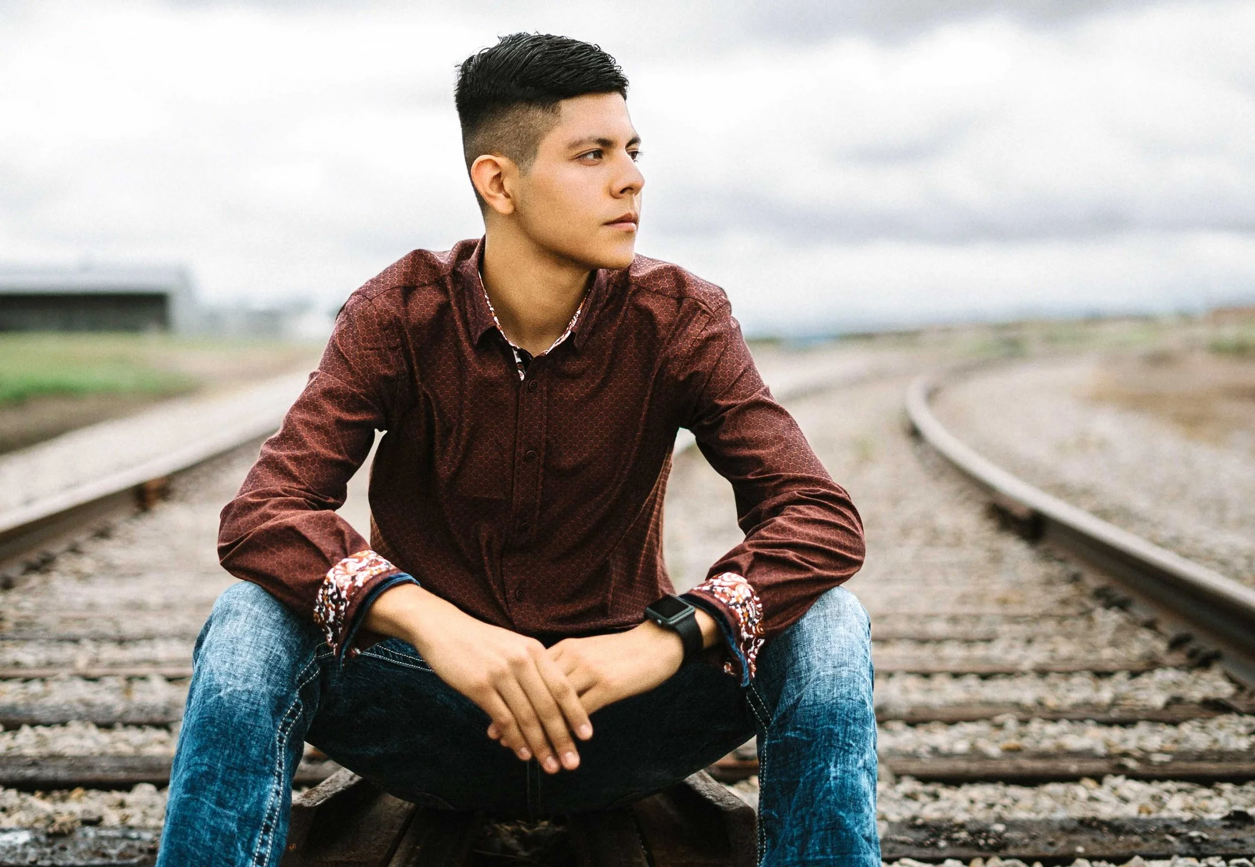 Young man sitting on railroad tracks, wearing a maroon shirt and jeans, looking to the side with a pensive expression.