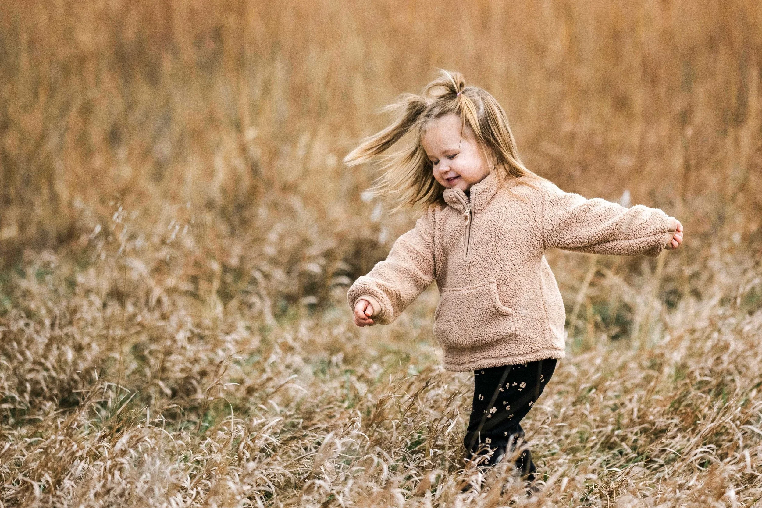 A young girl with blonde hair playing in a field of tall, dry grass, wearing a beige fleece jacket and black pants with a floral pattern.