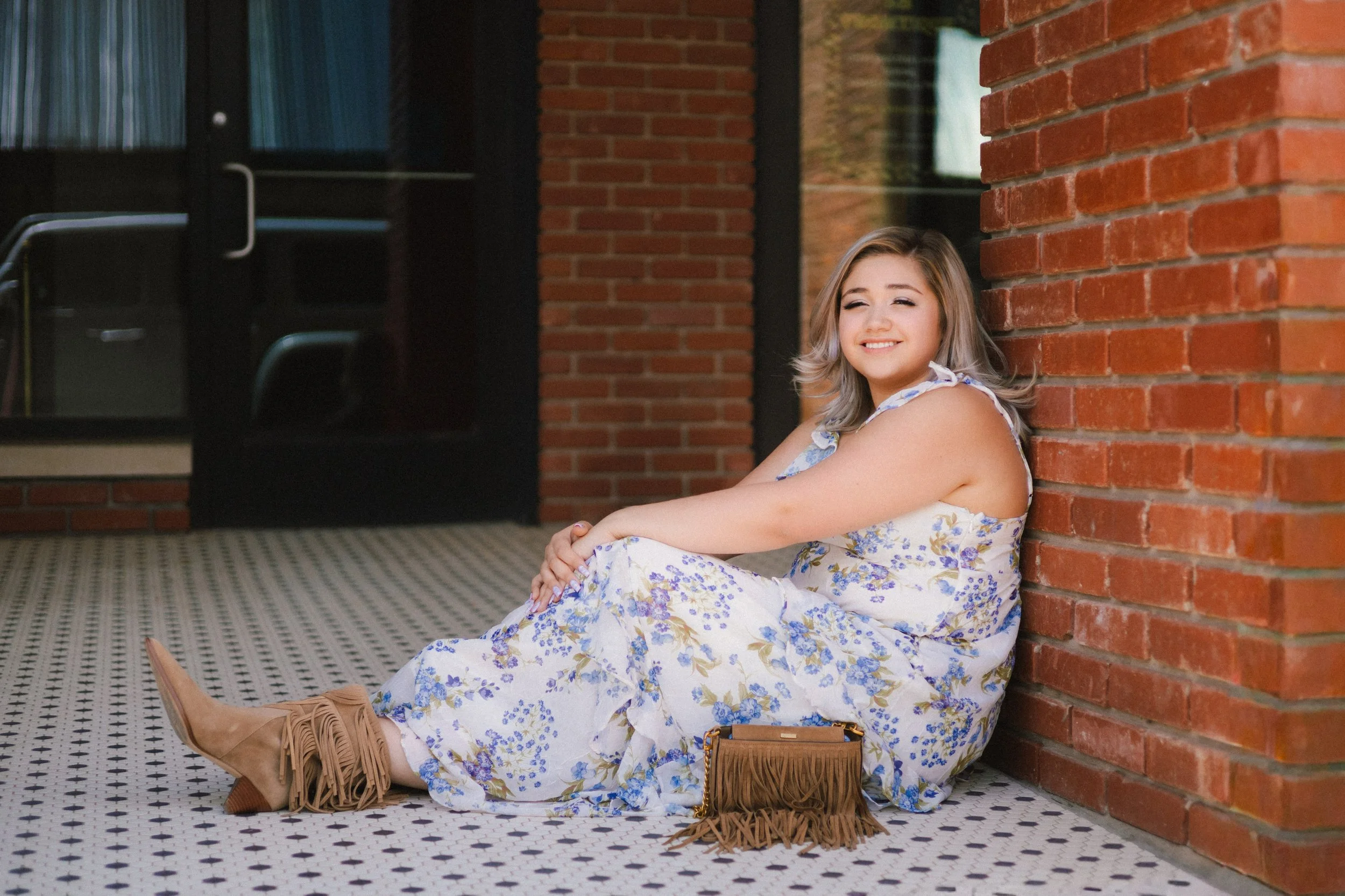 A smiling woman with blonde hair, wearing a floral dress, is sitting on the floor leaning against a brick wall outside a building. She has tan fringed boots and a tan purse beside her.