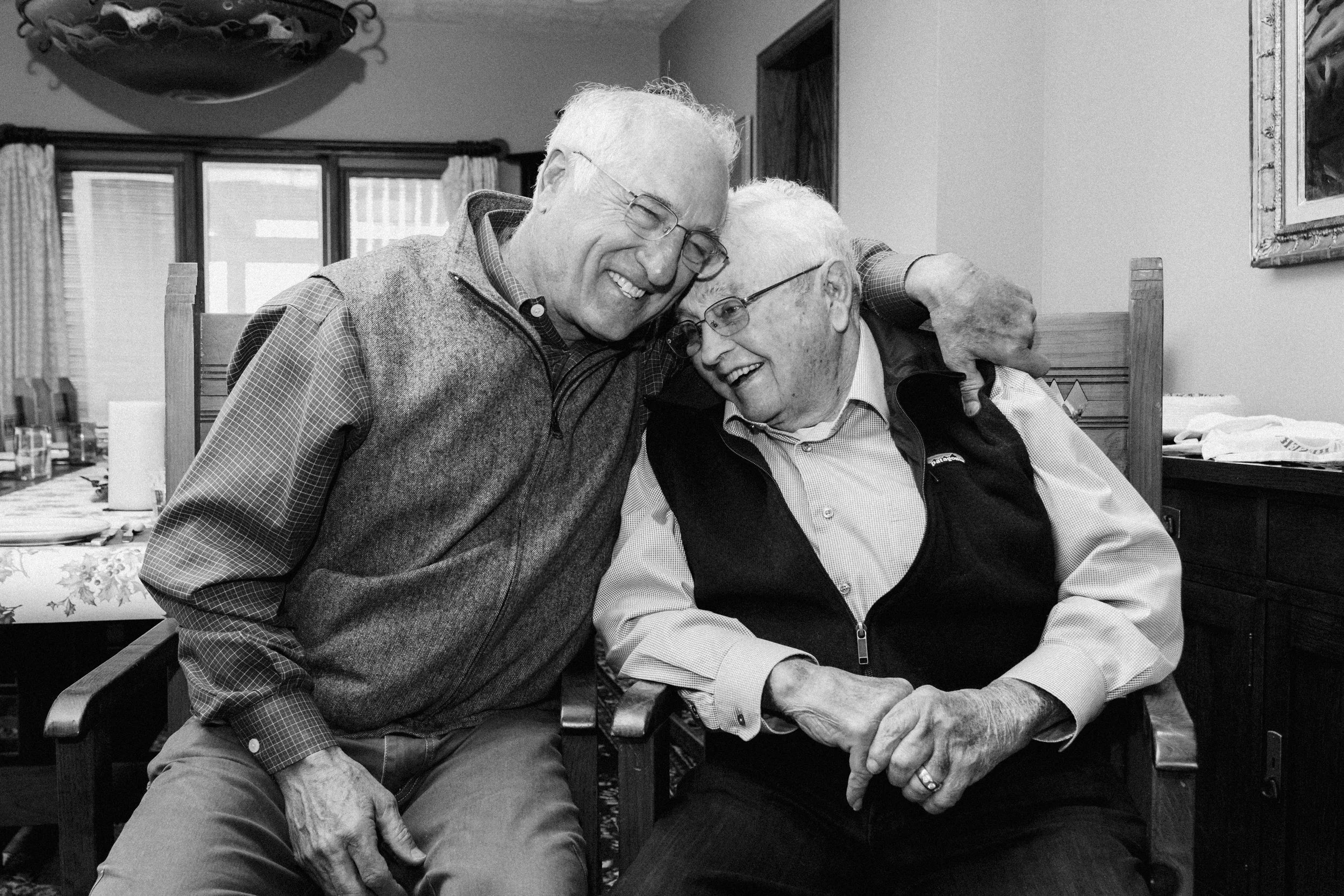Two elderly men sharing a tender, joyful moment, sitting close with one arm around each other's shoulders, smiling and leaning into each other in a cozy, homey room.