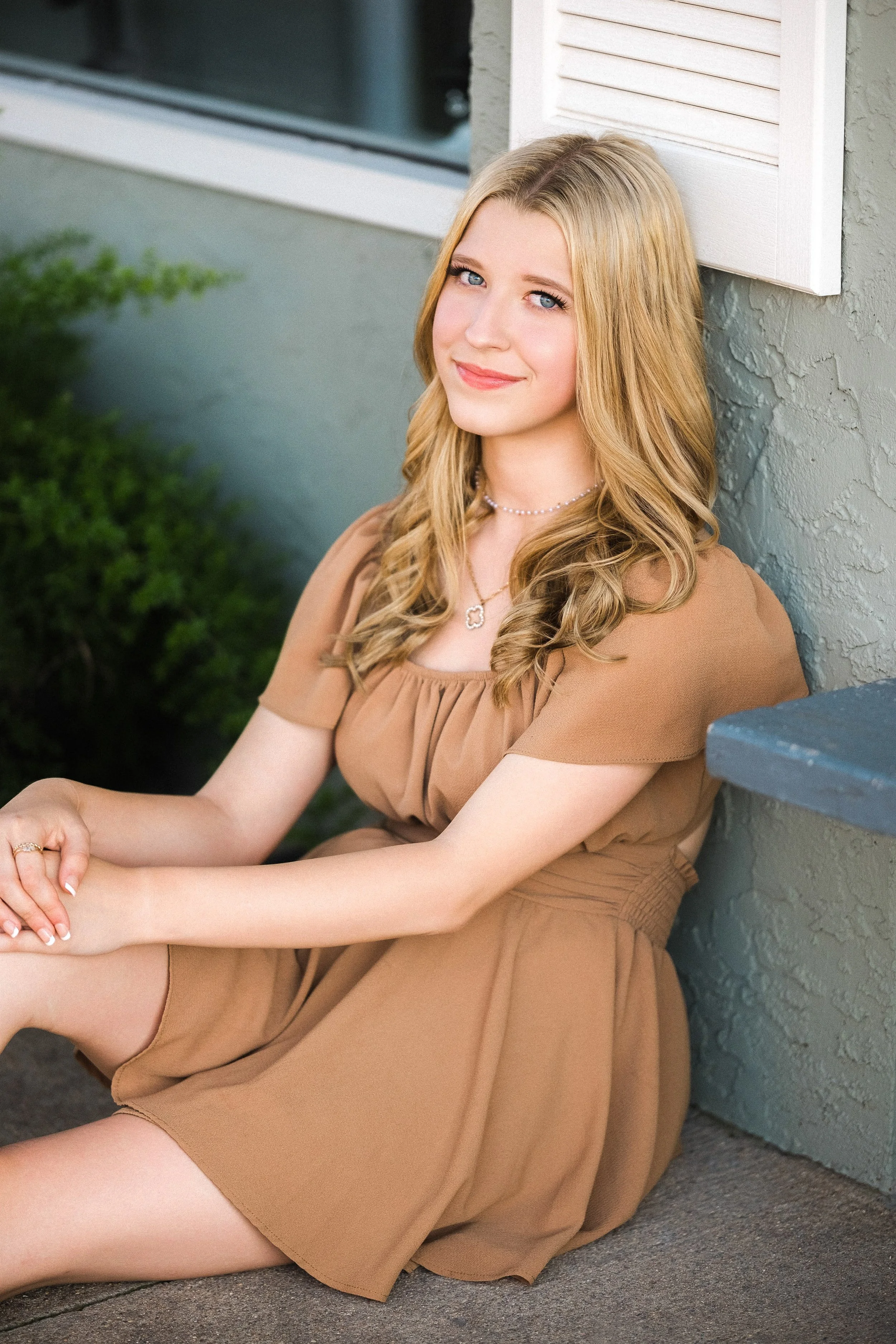 A young woman with long wavy blonde hair, blue eyes, and a light smile, sitting on the sidewalk against a textured wall, wearing a tan dress and jewelry, with greenery in the background.