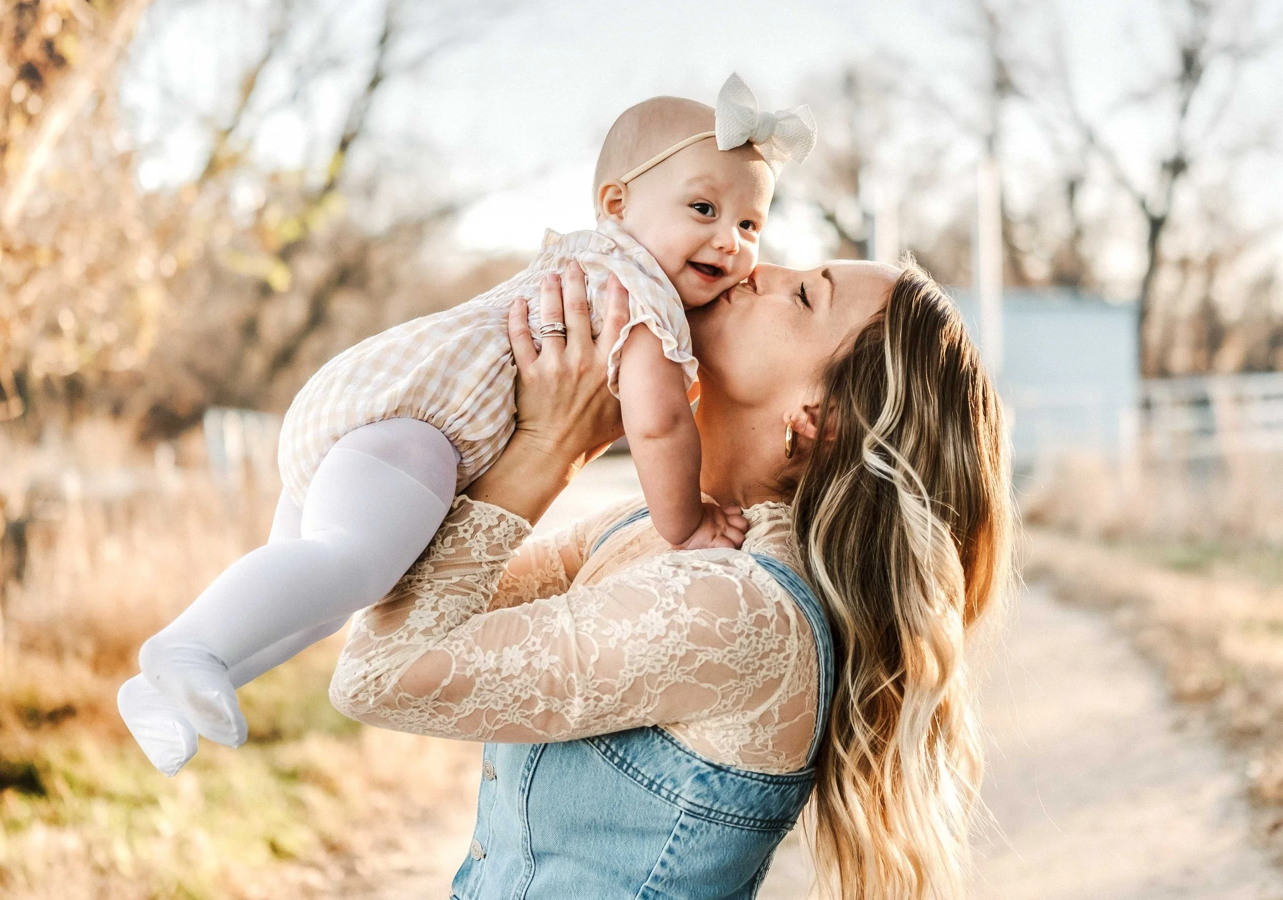 A woman with long wavy blonde hair, dressed in a lace top and denim vest, is holding a smiling baby girl in a plaid dress with white tights and a large white bow on her head; they are outdoors during autumn with trees and a pathway in the background.