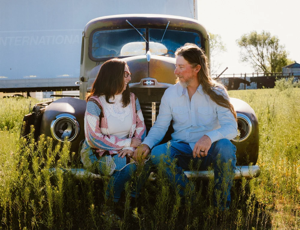 A couple sitting on the hood of a vintage truck in a grassy field