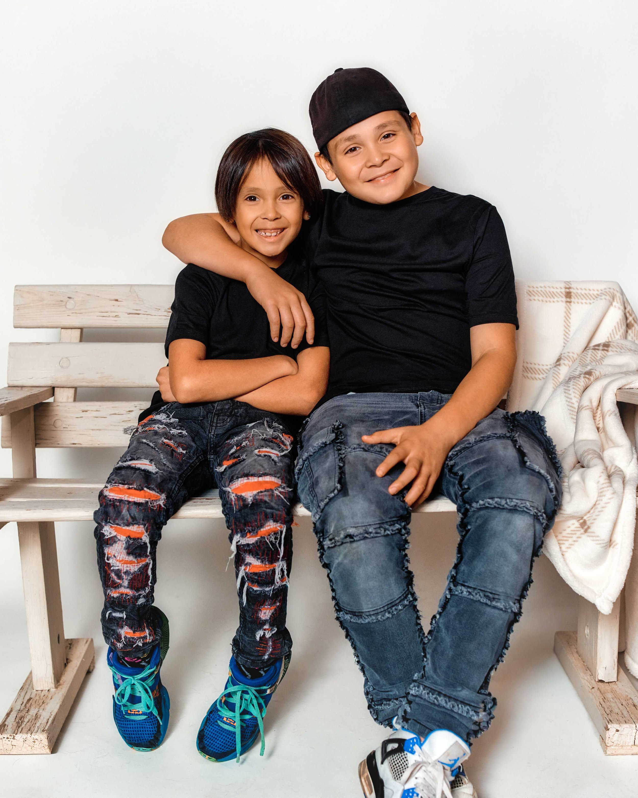 Two boys sitting on a wooden bench, smiling, one boy has his arm around the other.
