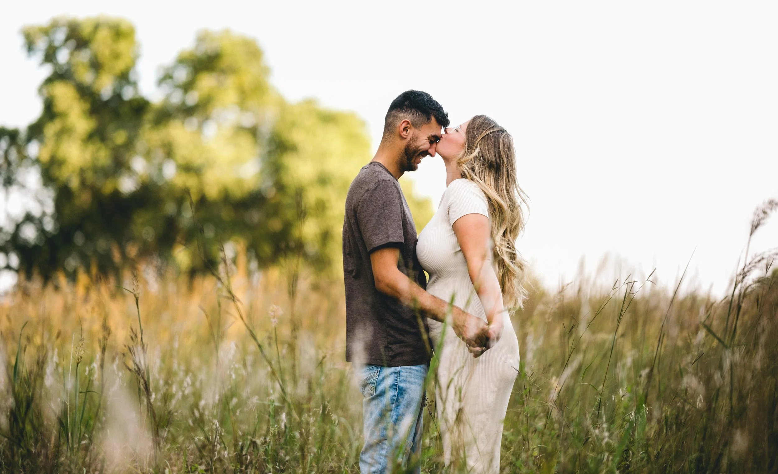 A couple standing in a grassy field with trees in the background, sharing a kiss and holding hands, during the daytime.