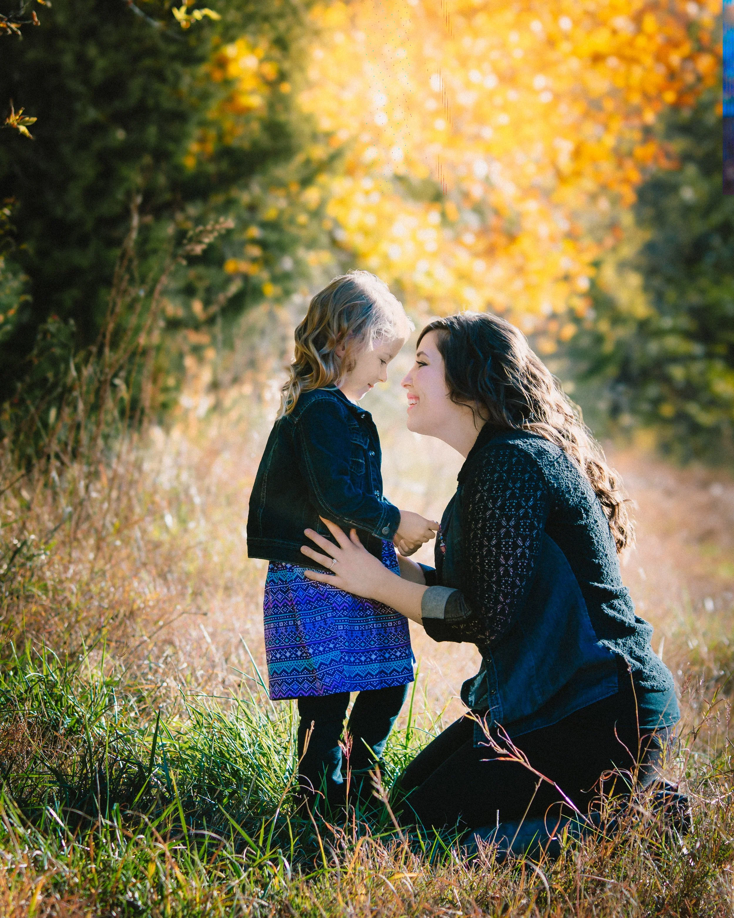 A woman kneeling in a grassy field, holding hands with a young girl, both smiling and touching foreheads in a sunny, autumnal outdoor setting.